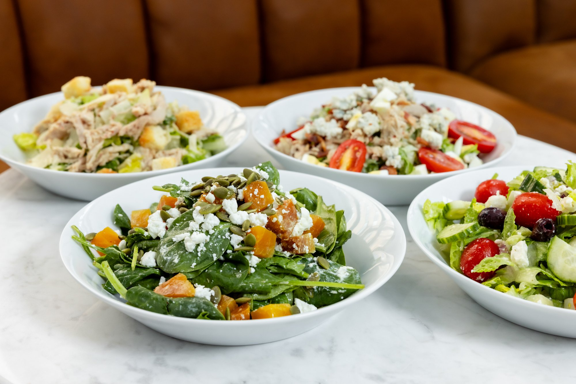 Four white bowls of salad on a white marble table, with a brown cushioned seat in the background. The salads include various vegetables, cheese, and other toppings.