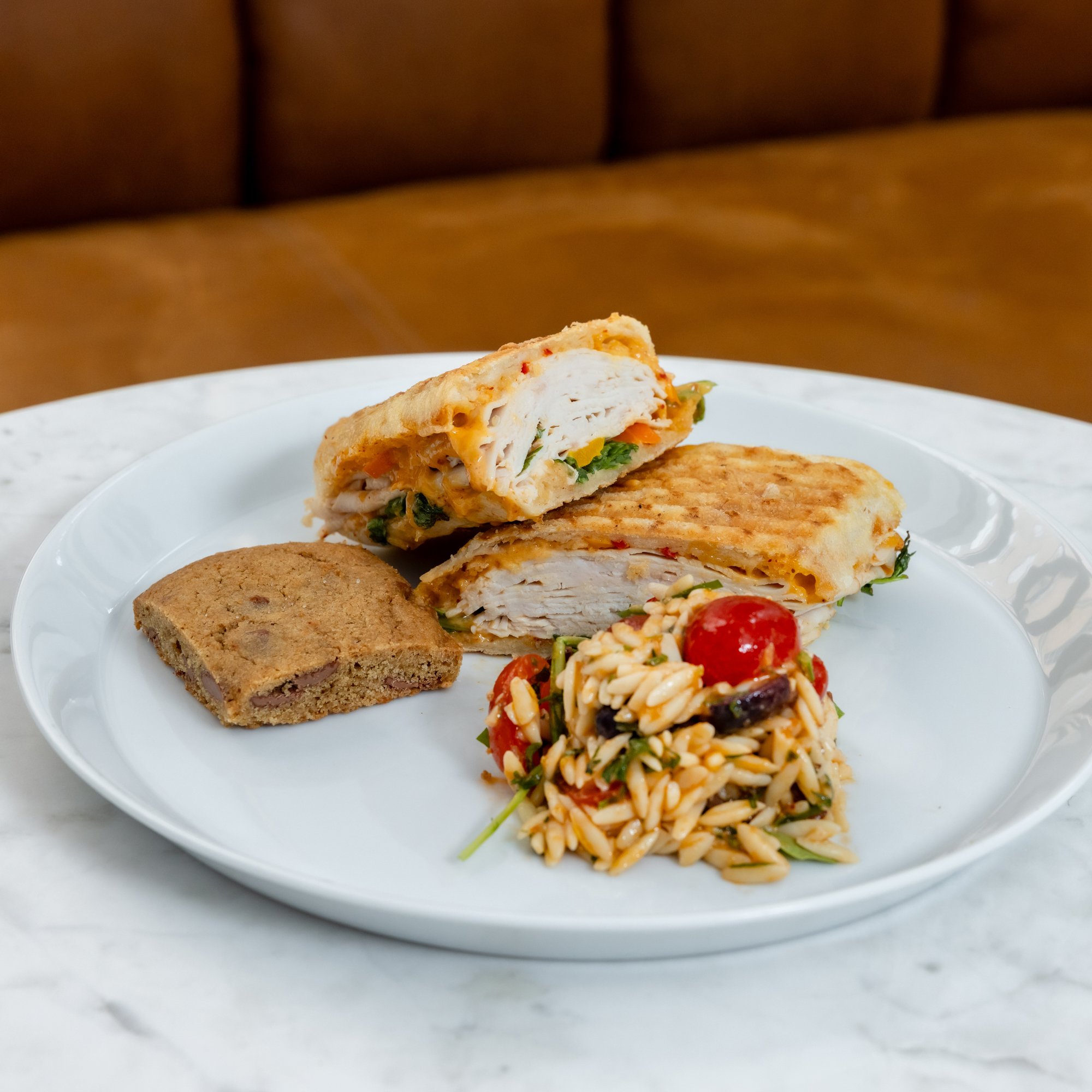 A white plate with grilled chicken sandwich, a piece of cookie, and a serving of pasta salad with cherry tomatoes on a marble table, with a brown cushioned bench in the background.