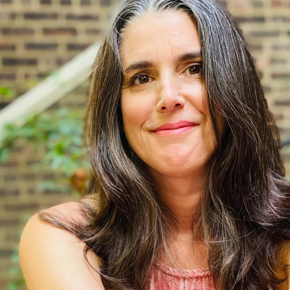 Alicia Nosenzo- A woman with long, dark, wavy hair smiling slightly in front of a brick wall with some greenery.