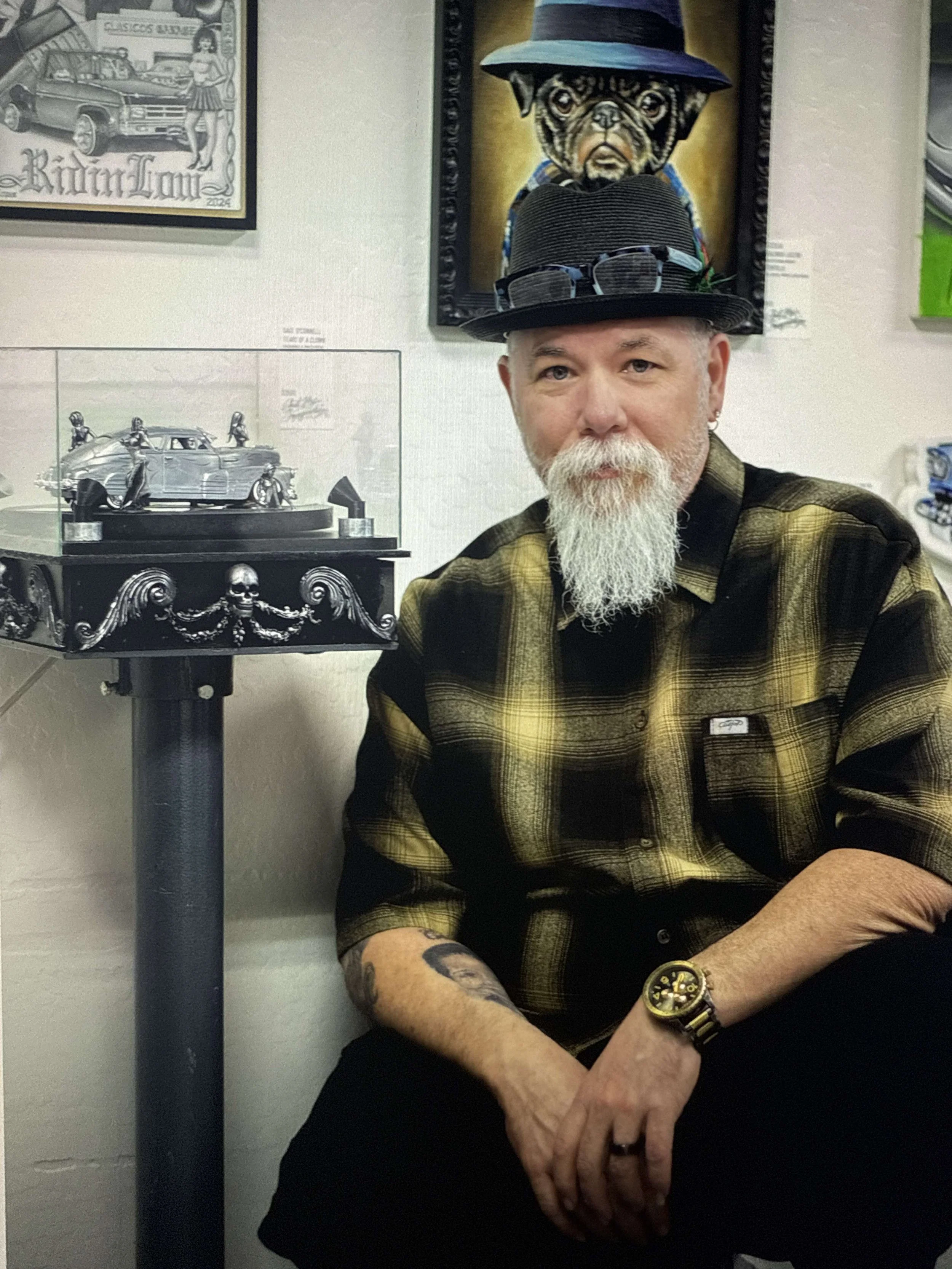 Sage , a man with a white beard, wearing a yellow and black plaid shirt, black hat with sunglasses on top, and a watch, is sitting next to a display case with a model car. There are framed artworks of cars and a dog on the wall behind him.