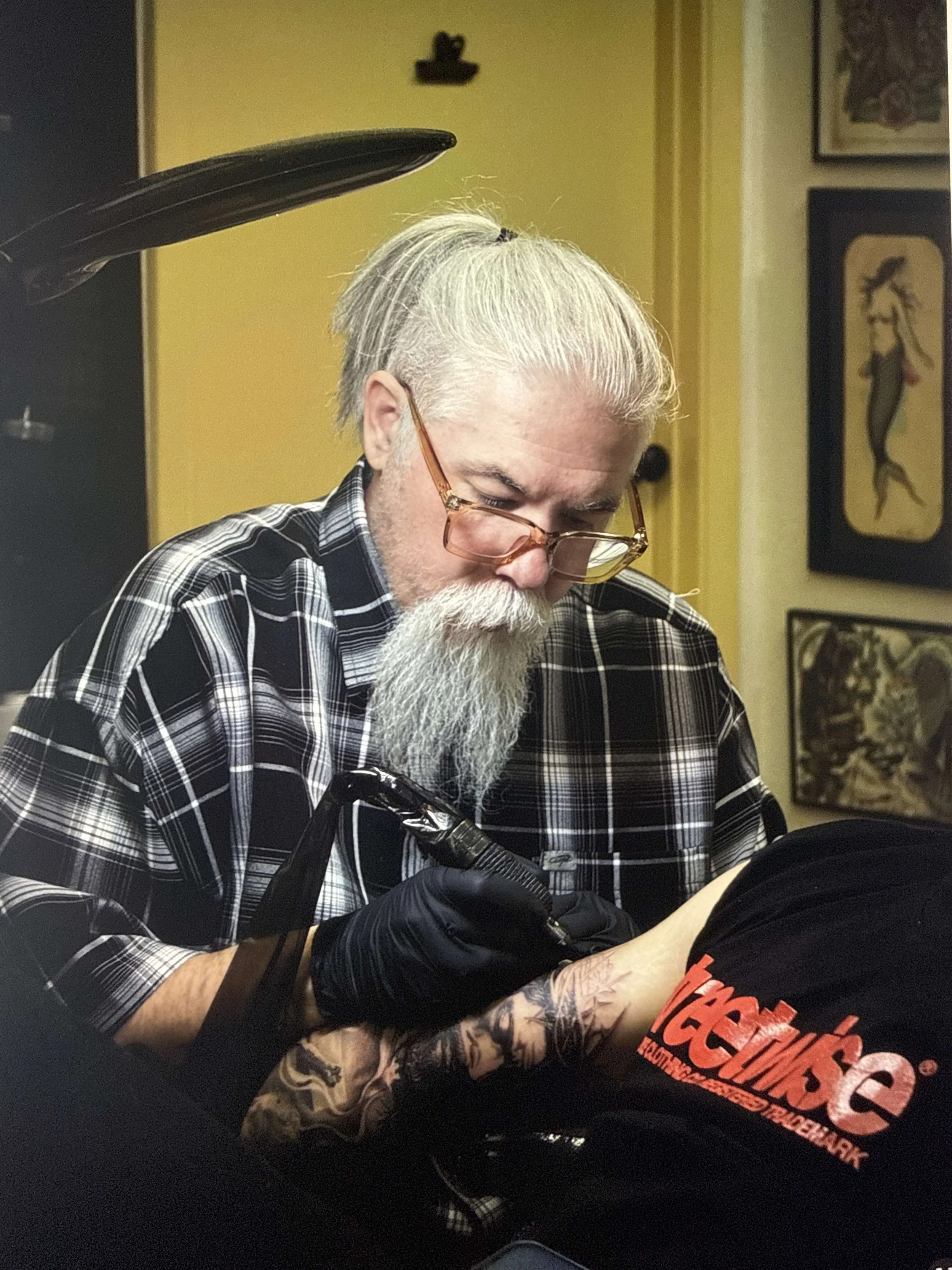 Sage, a man with white hair and a beard, wearing glasses and a black plaid shirt, is getting a tattoo on his arm. He is using a tattooing machine, and the tattoo features detailed artwork. The background includes framed artwork and a yellow wall.