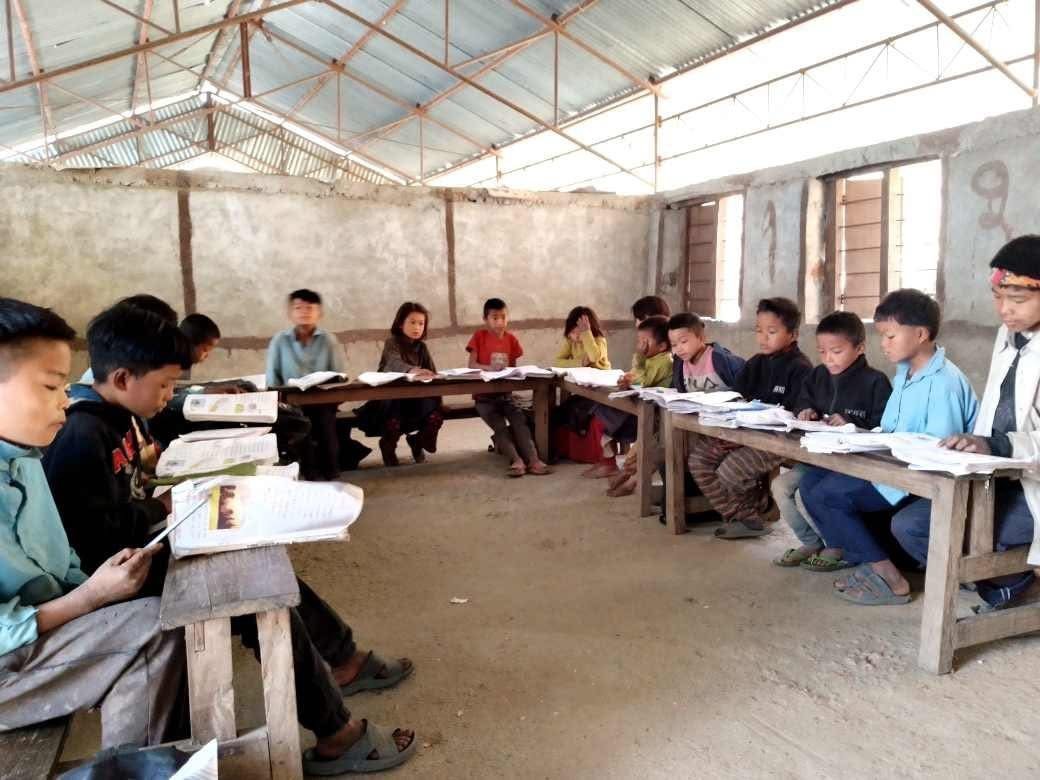 A group of Nepalese students seated in a simple classroom, arranged in a semicircle. They are reading from textbooks placed on rows of wooden desks. The classroom has a metal roof, concrete walls, and several ventilation windows. 