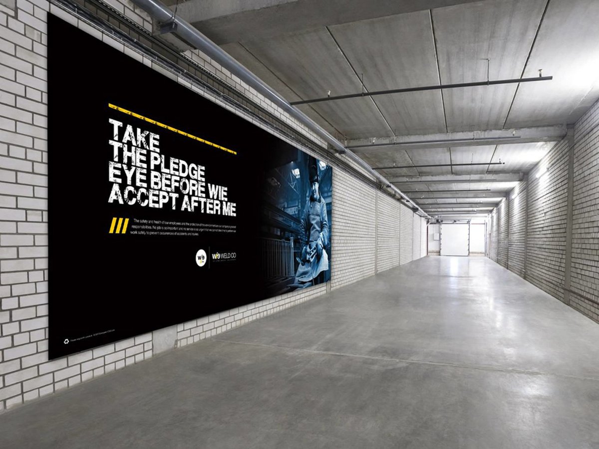 Industrial corridor with a safety pledge poster on a brick wall. The poster reads "Take the pledge, eye before we accept after me" in bold text with industrial imagery. The corridor has a concrete ceiling and floor, with fluorescent lighting.