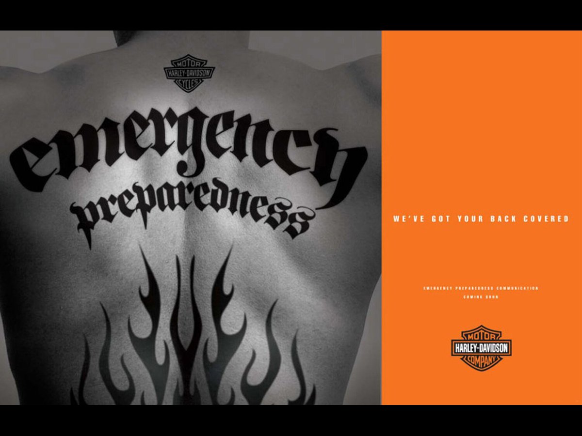 Close-up of a tattooed human back featuring "emergency preparedness" in stylized script, a Harley-Davidson logo at the top, and black flame designs at the bottom. Orange panel on the right says "WE'VE GOT YOUR BACK COVERED" and includes another Harle