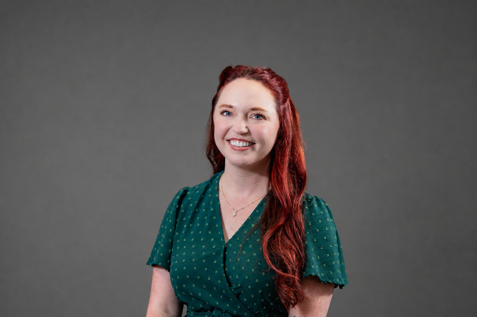 Portrait of a woman with long, wavy, red hair wearing a green blouse with small gold dots, standing in front of a white brick wall, smiling.