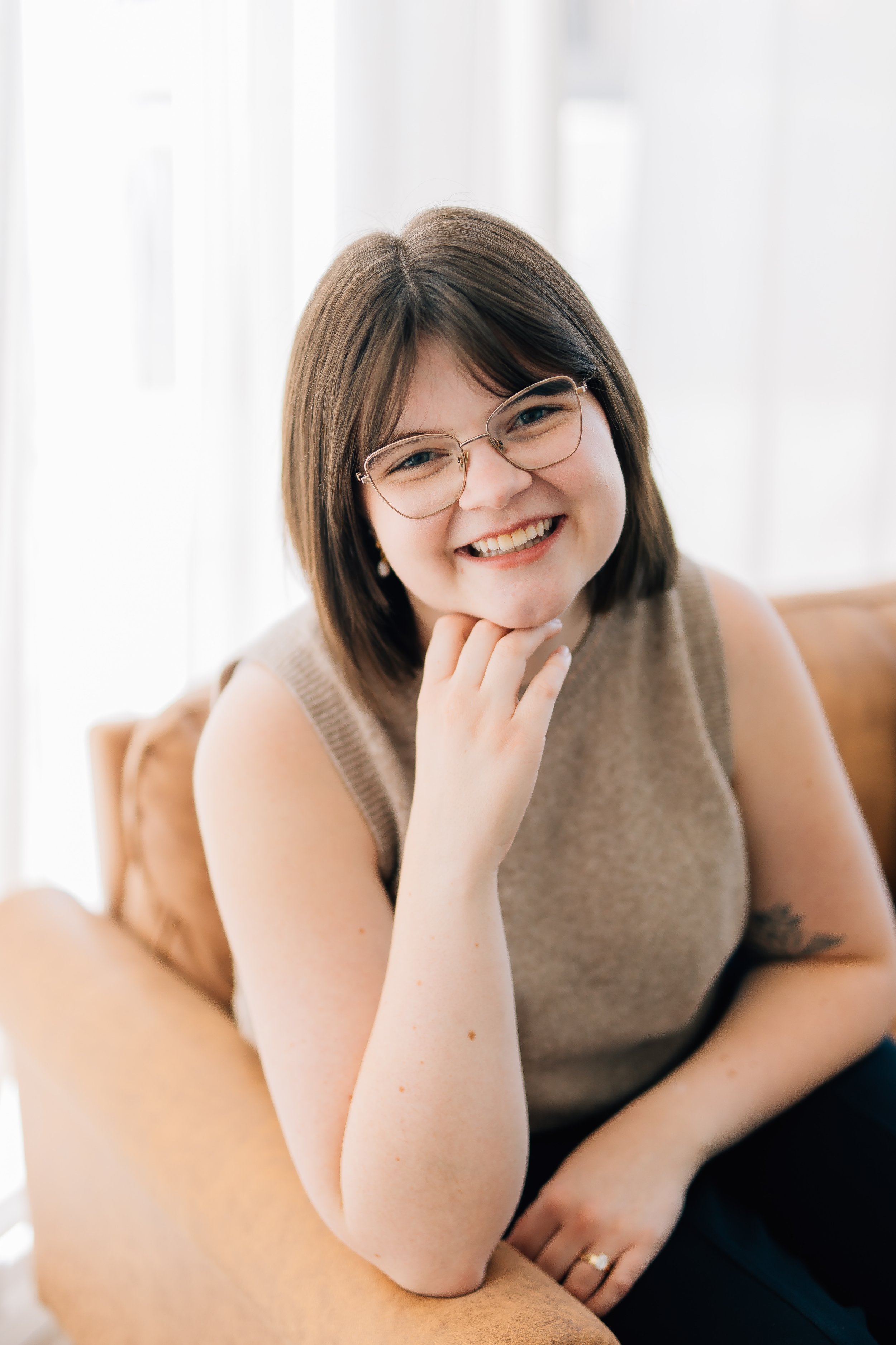 A smiling woman with glasses and brown hair, wearing a beige sleeveless top, resting her chin on her hand, sitting closely with a person with visible arm, in a bright room with white curtains.