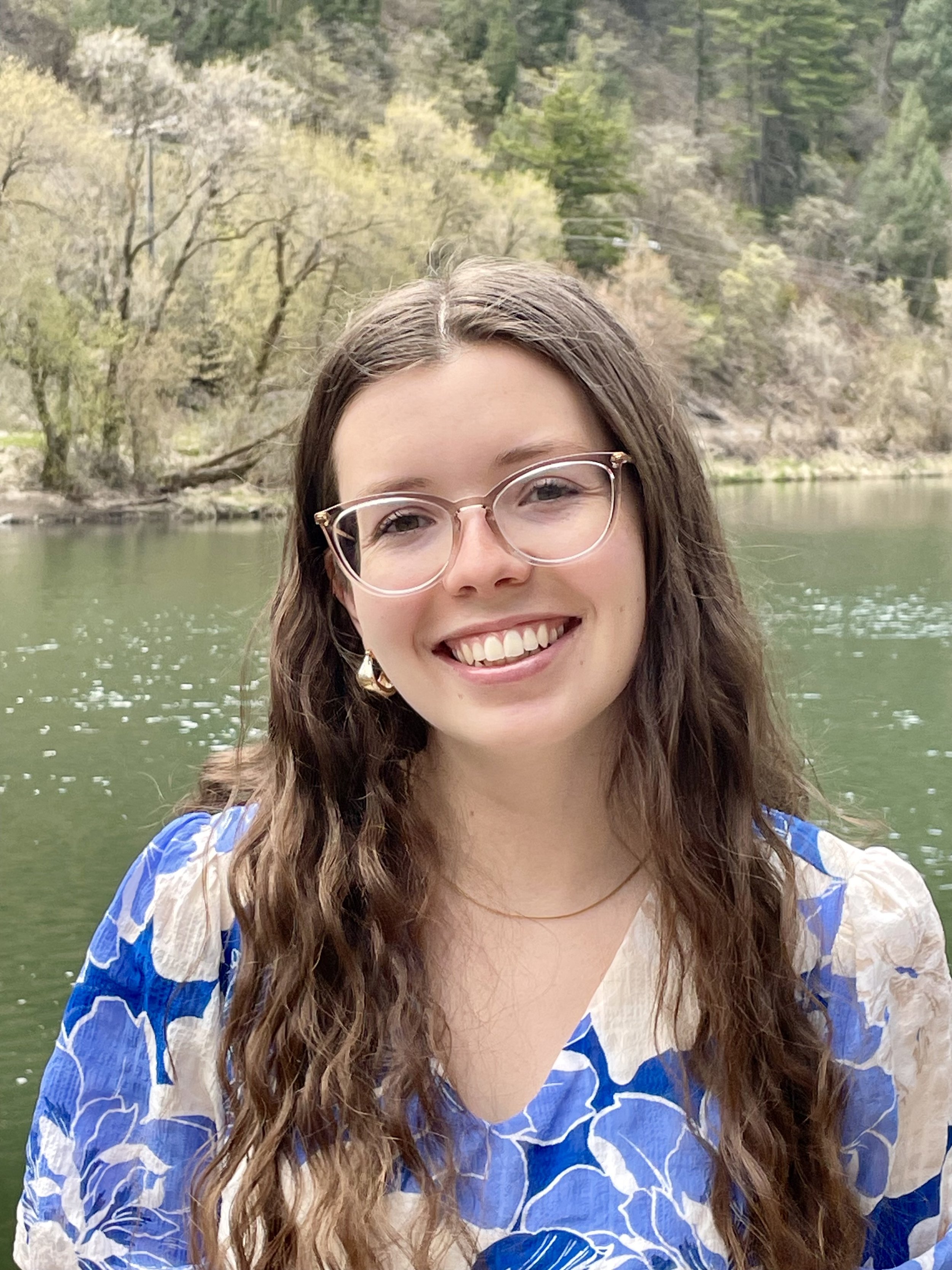 A young woman with long wavy brown hair, wearing glasses and a colorful dress with blue and white patterns, smiling outdoors near a body of water with a forested background.