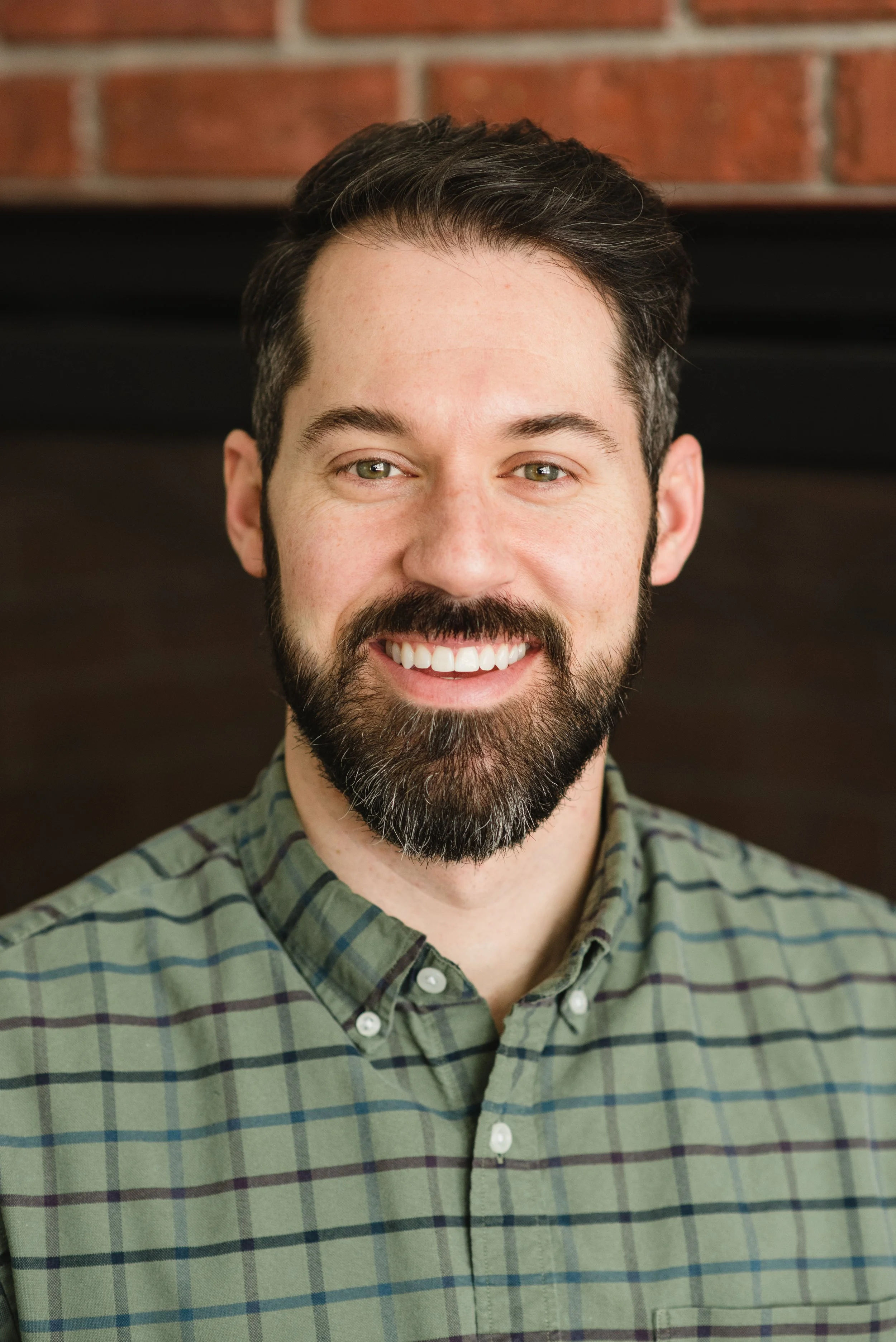 Close-up of a smiling man with dark hair, beard, and mustache, wearing a green and black checkered button-up shirt, standing against a brick wall background.