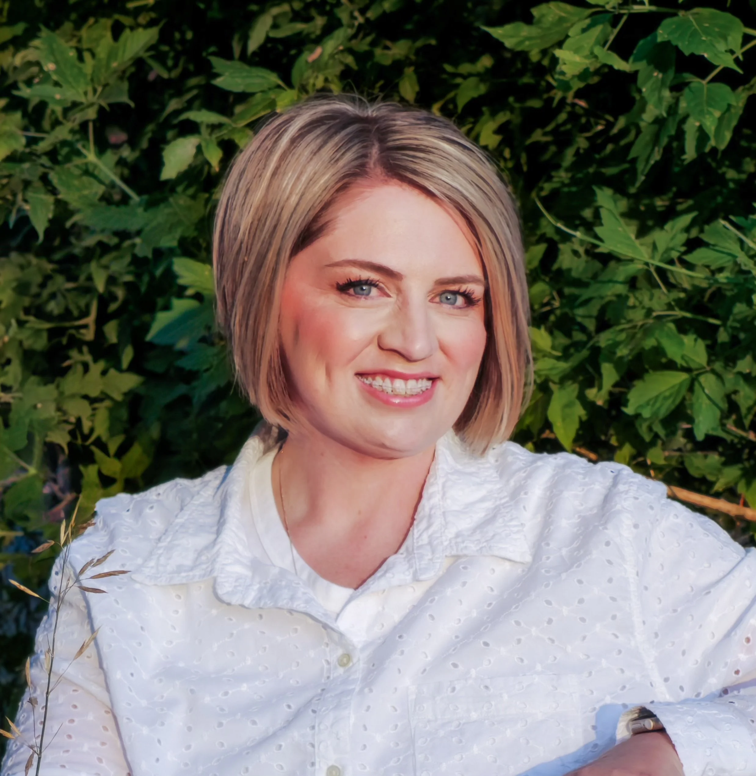 A woman with blonde hair and blue eyes smiling outdoors in front of green leafy bushes, wearing a white dress with textured fabric and a white bracelet on her wrist.
