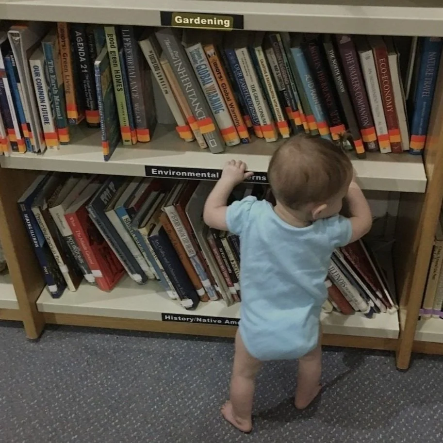 Small baby in a light blue onesie browses library shelf of books on gardening, environmental concerns, natural history, and Native American practices.
