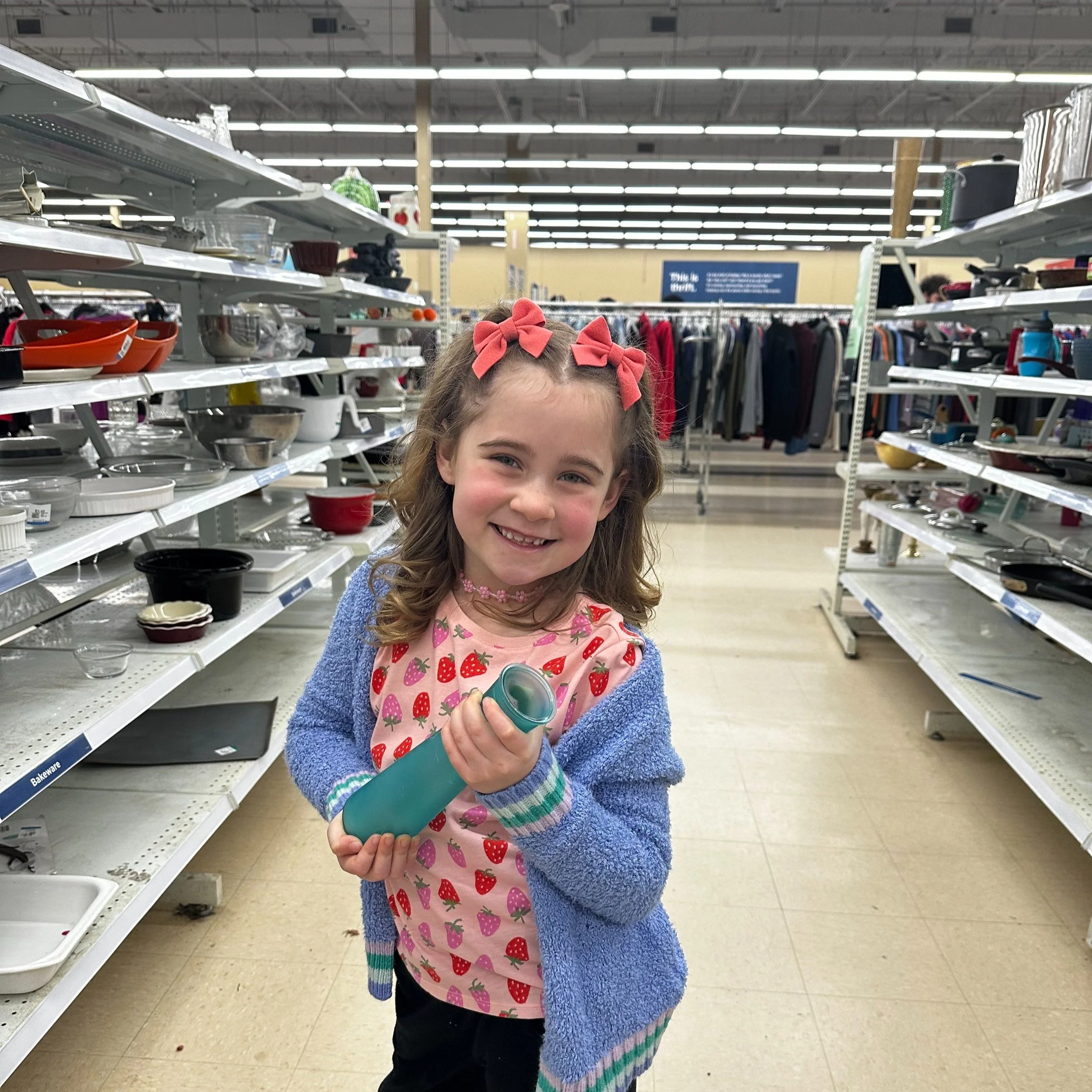 Little girl with strawberry shirt and purple cardigan joyfully posing with flower vase between thrift store aisles. She appears to be enjoying her experience at the second hand store.