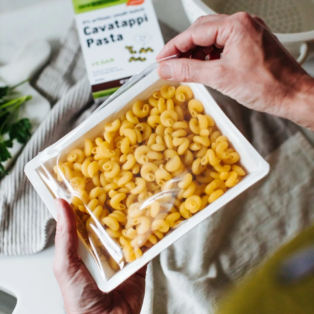 Image of hand holding package of pasta in recyclable container, opening the plastic top to get to the pasta.