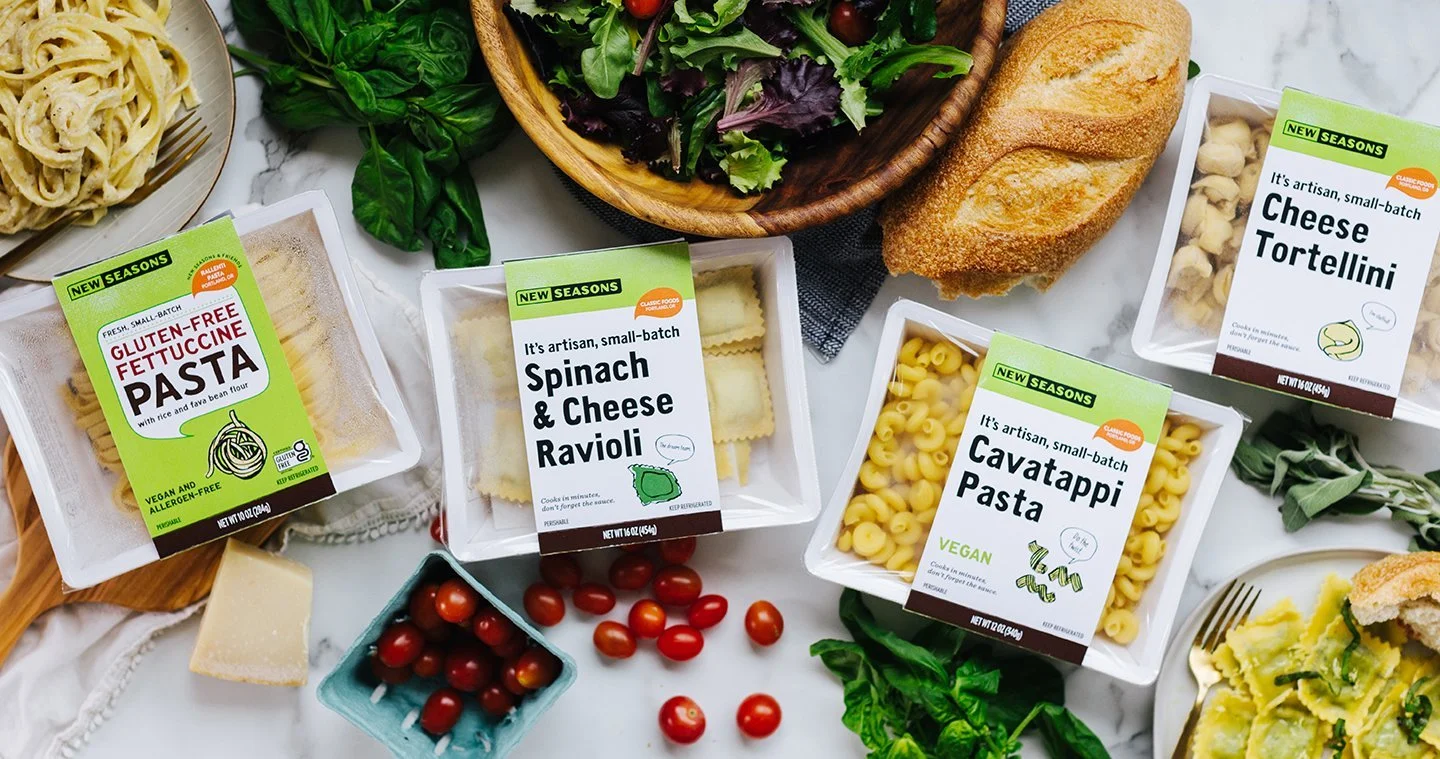 Image of three different packaged fresh pastas, a bowl of greens, cherry tomatoes, basil and fresh cooked pasta.
