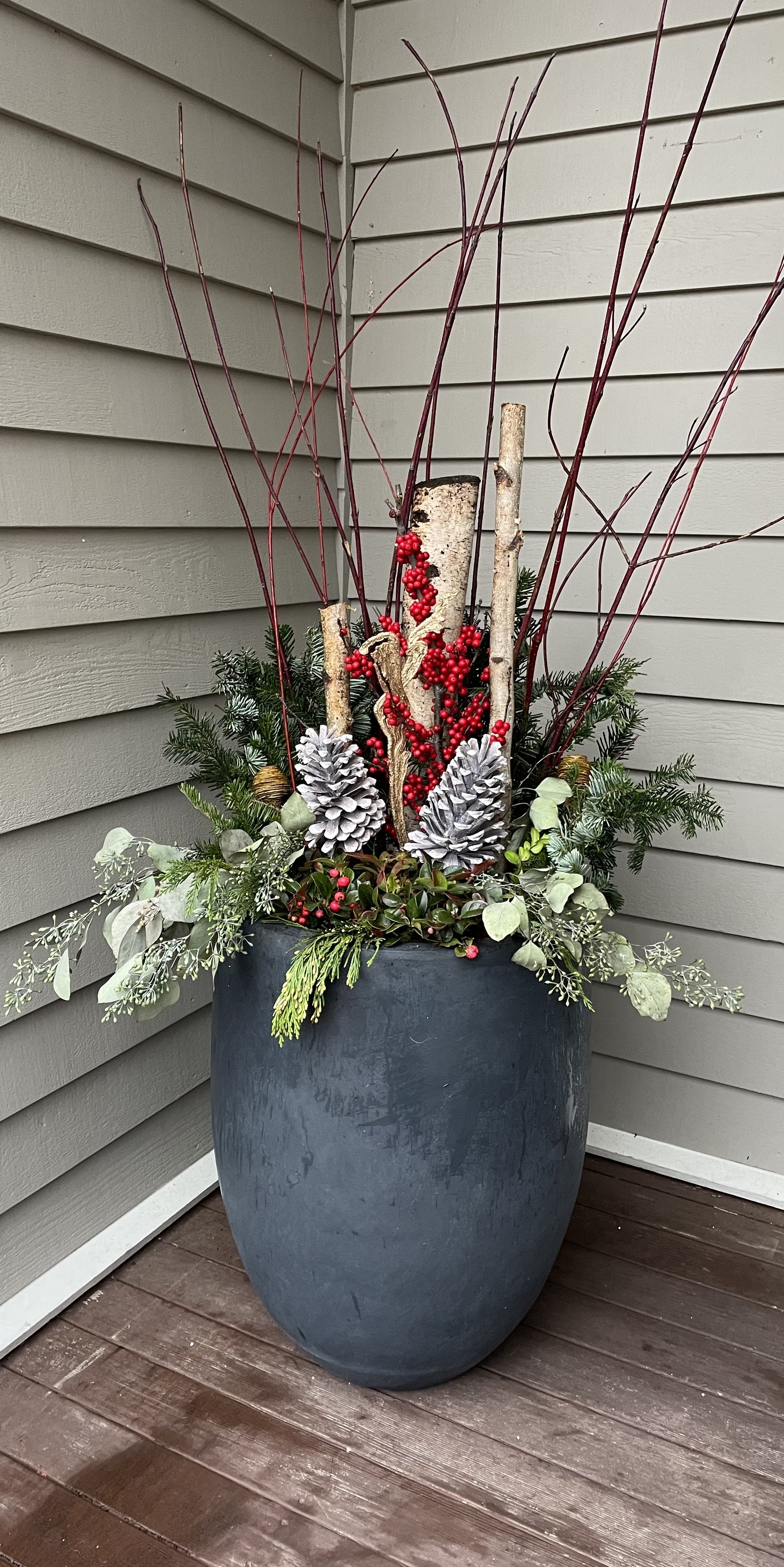 Outdoor decorative planter with birch logs, red berries, pinecones, and evergreen branches.