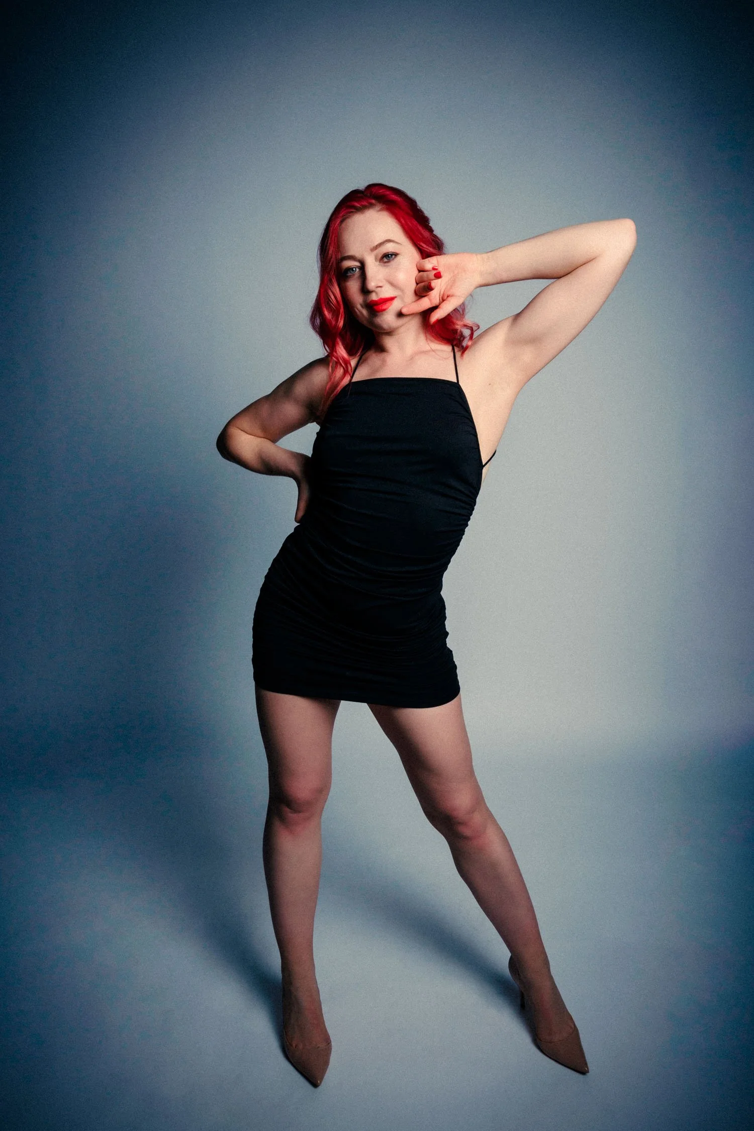 Femme aux cheveux rouges portant une robe noire, faisant une pose de model en studio.