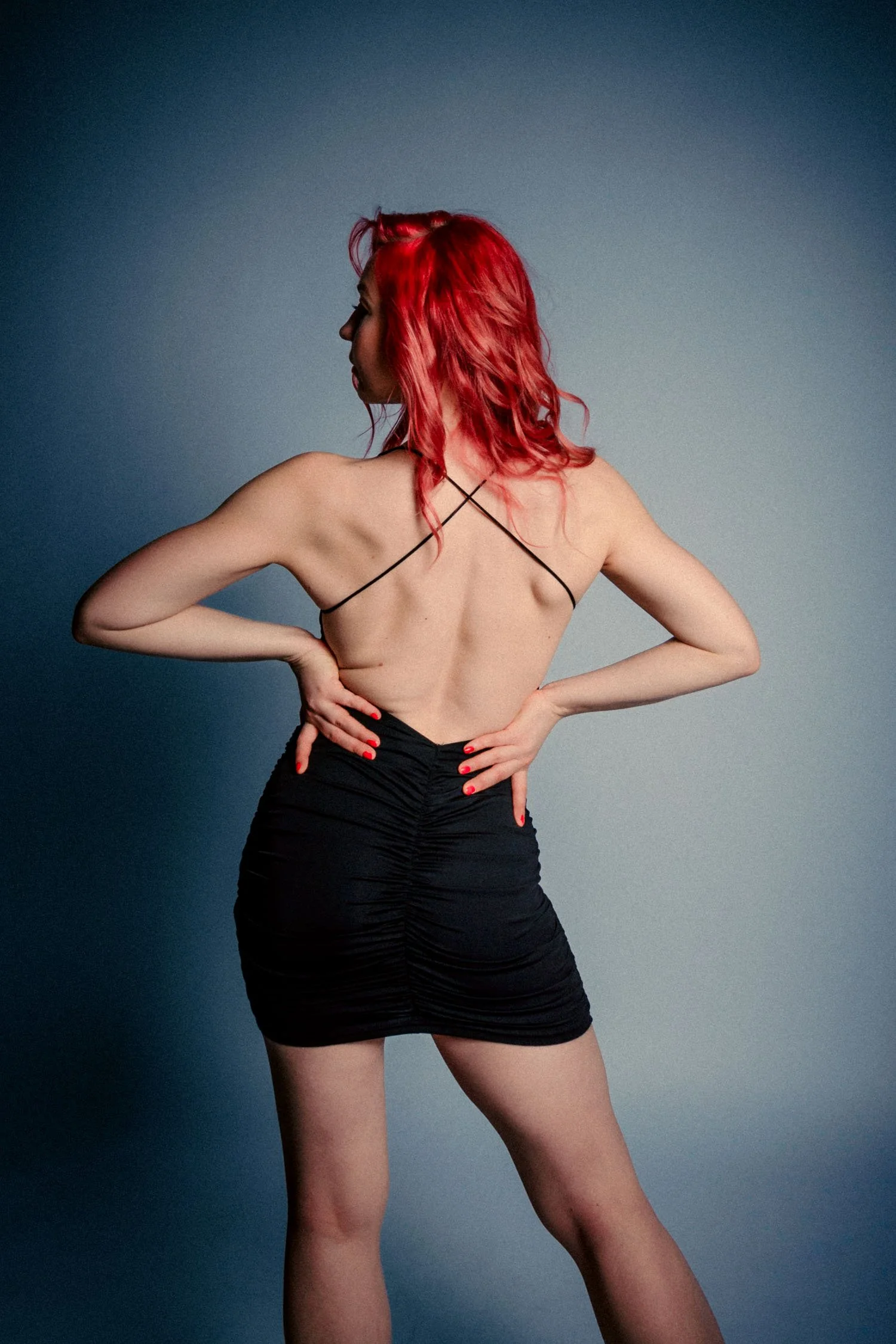 Une femme avec des cheveux rouges, portant une robe noire sexy avec un dos nu, posant avec les mains sur les hanches devant un fond gris