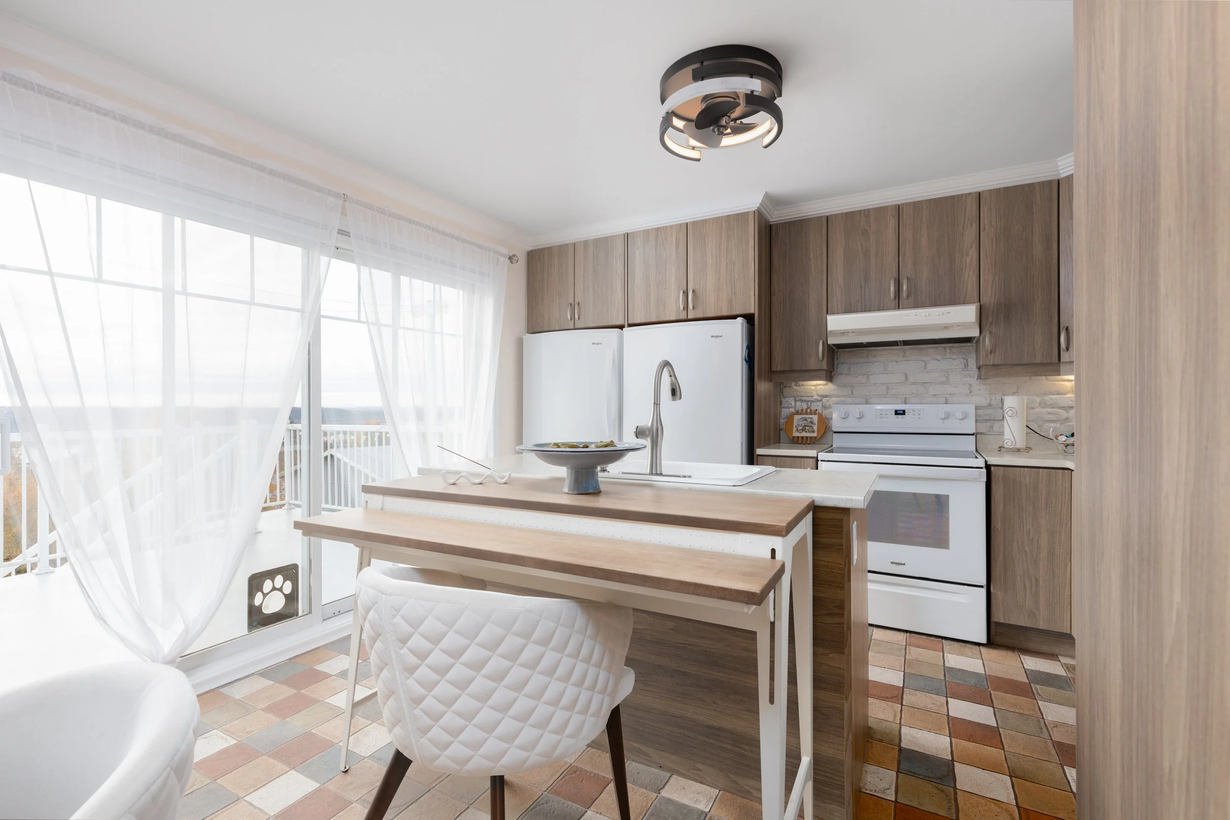 Cuisine moderne avec fenêtres grandes, rideaux en voile blanc, meubles en bois, plan de travail avec évier, cuisinière blanche, réfrigérateur blanc, plafonnier design, sol en carreaux de terre cuite, décor simple.