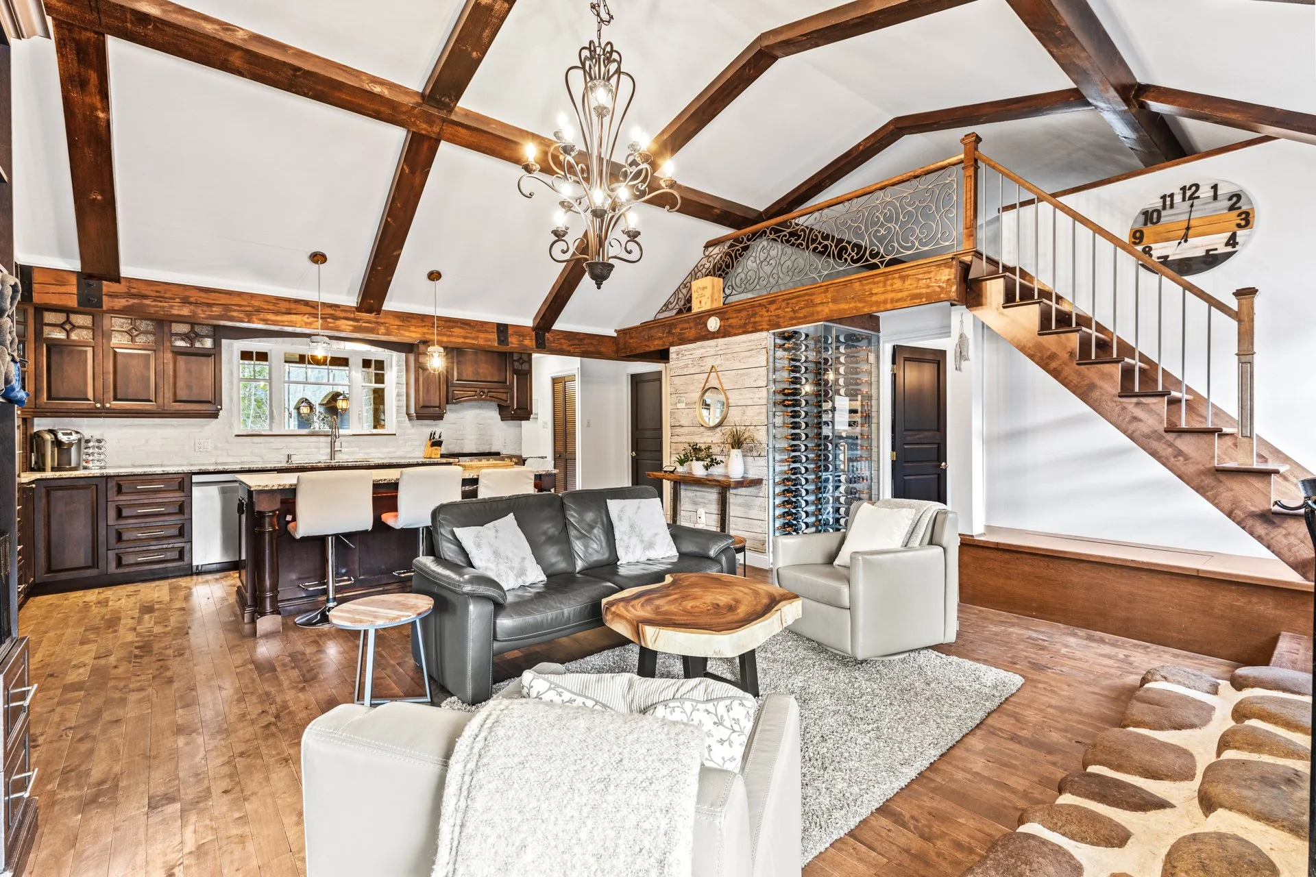 Salon avec sofa, fauteuils, cheminée en bois, escalier en bois menant à un espace mezzanine, mur en bois clair, murs blancs, horloge murale, étagère à vin, luminaires suspendus et chandelier sculptural, décoration intérieure chaleureuse.