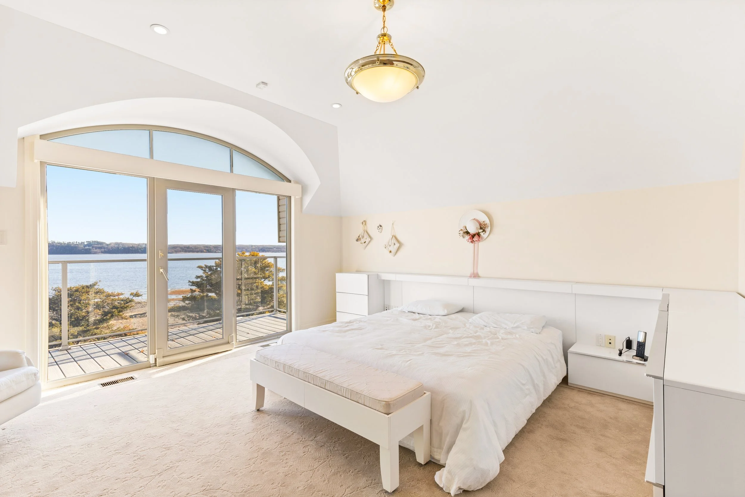 Chambre lumineuse avec un lit king-size, vue sur un lac à travers une grande fenêtre en forme d'arc, décoration minimaliste avec un plafond en pente, et un chandelier suspendu.