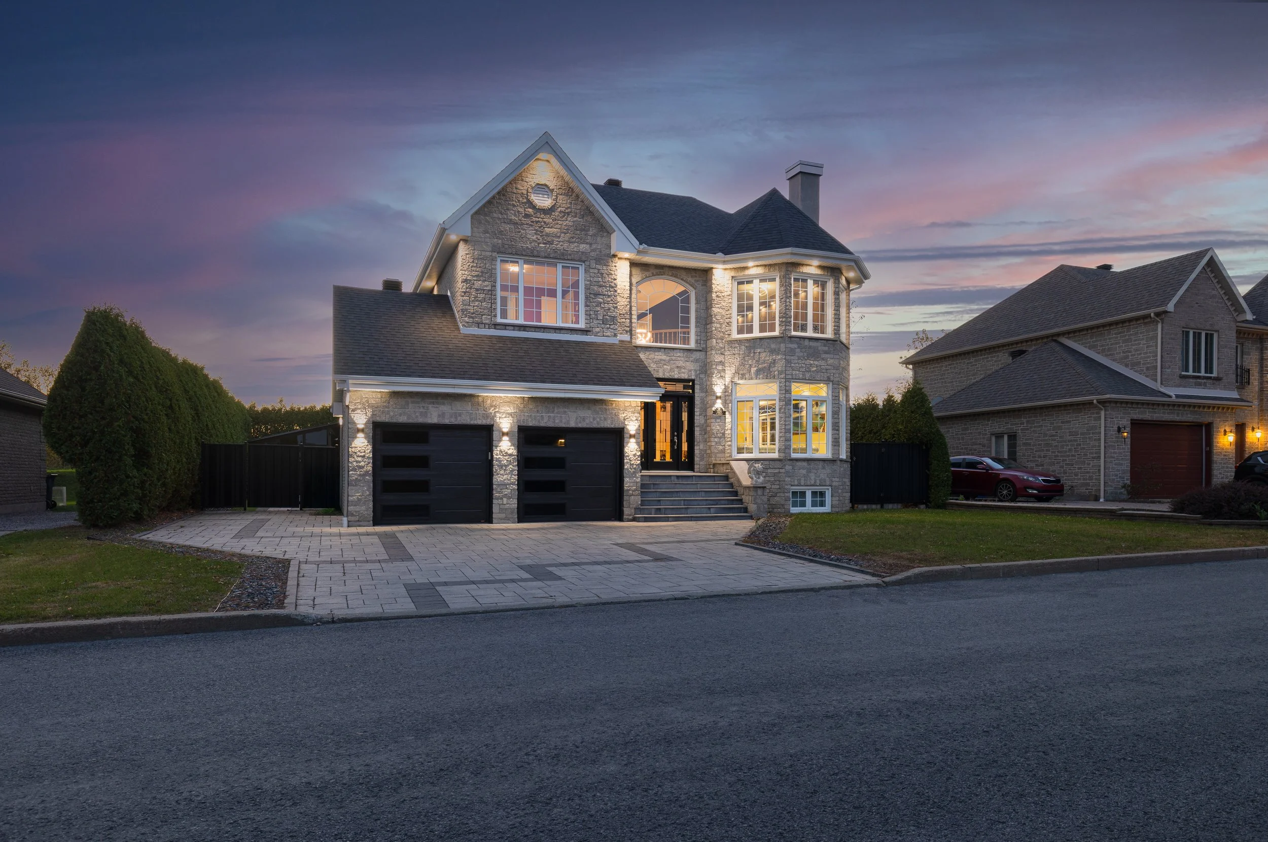 Une grande maison en pierre avec un garage double, fenêtres éclairées, entourée d'une pelouse et un chemin en pavés, sous un ciel au coucher du soleil.
