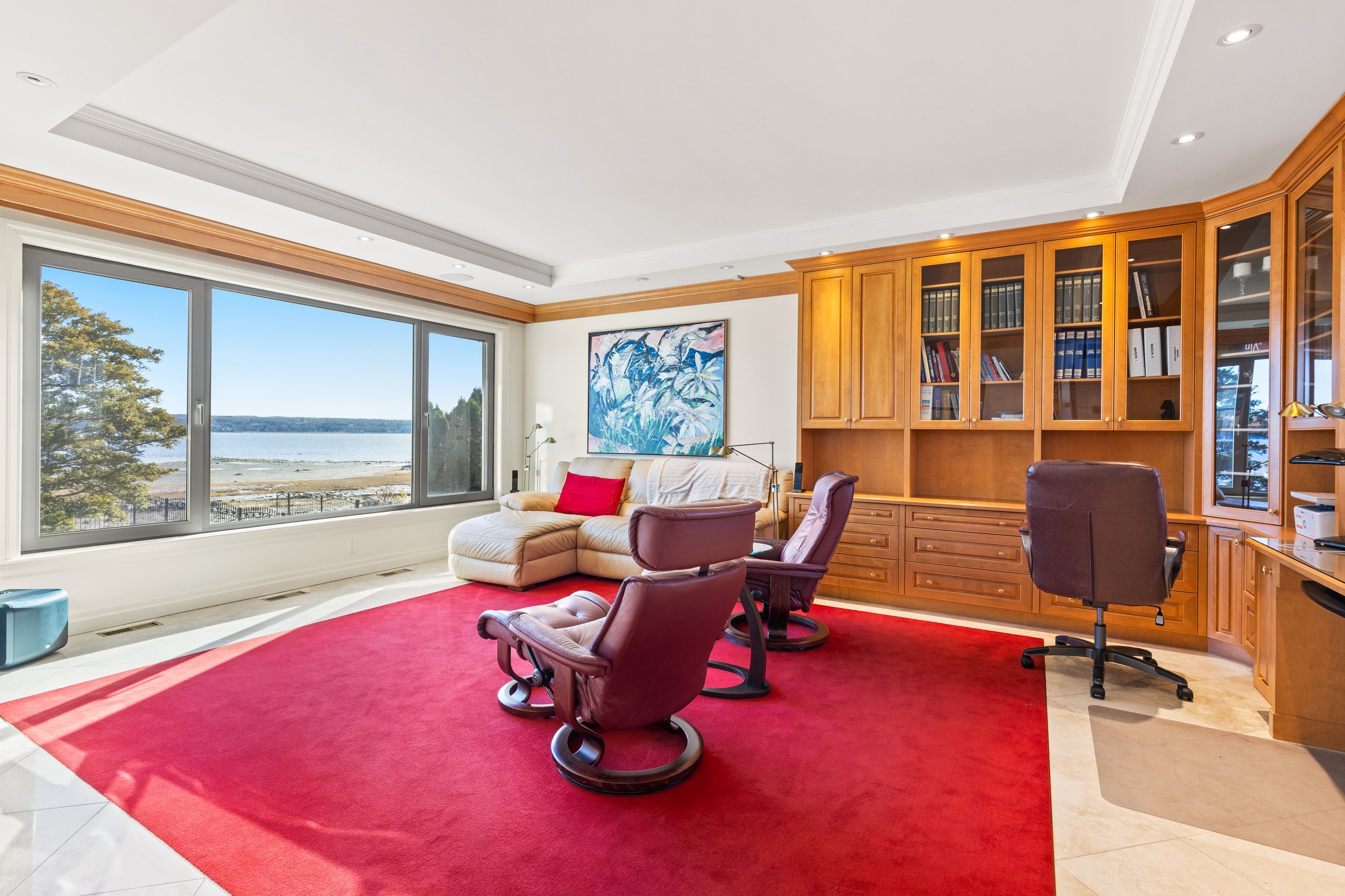 Salon avec grande fenêtre offrant vue sur un lac, comprenant un canapé beige, des fauteuils en cuir marron, un tapis rouge et une bibliothèque en bois