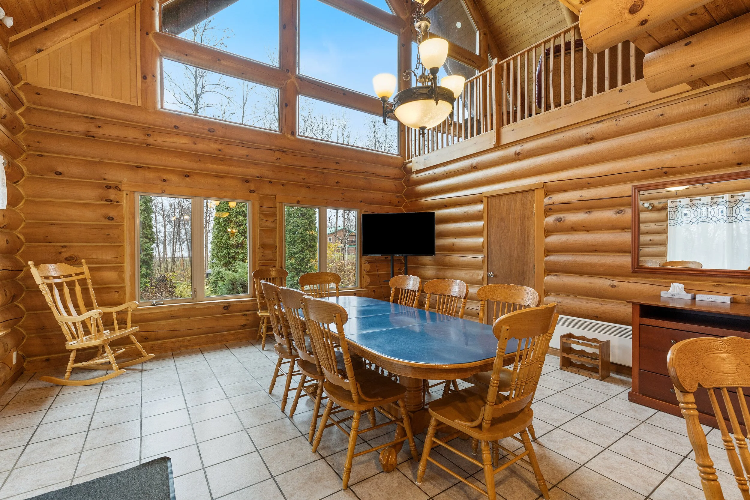 Salle à manger en bois rond avec table en bois, chaises et grand chandelier, fenêtres offrant vue sur des arbres.