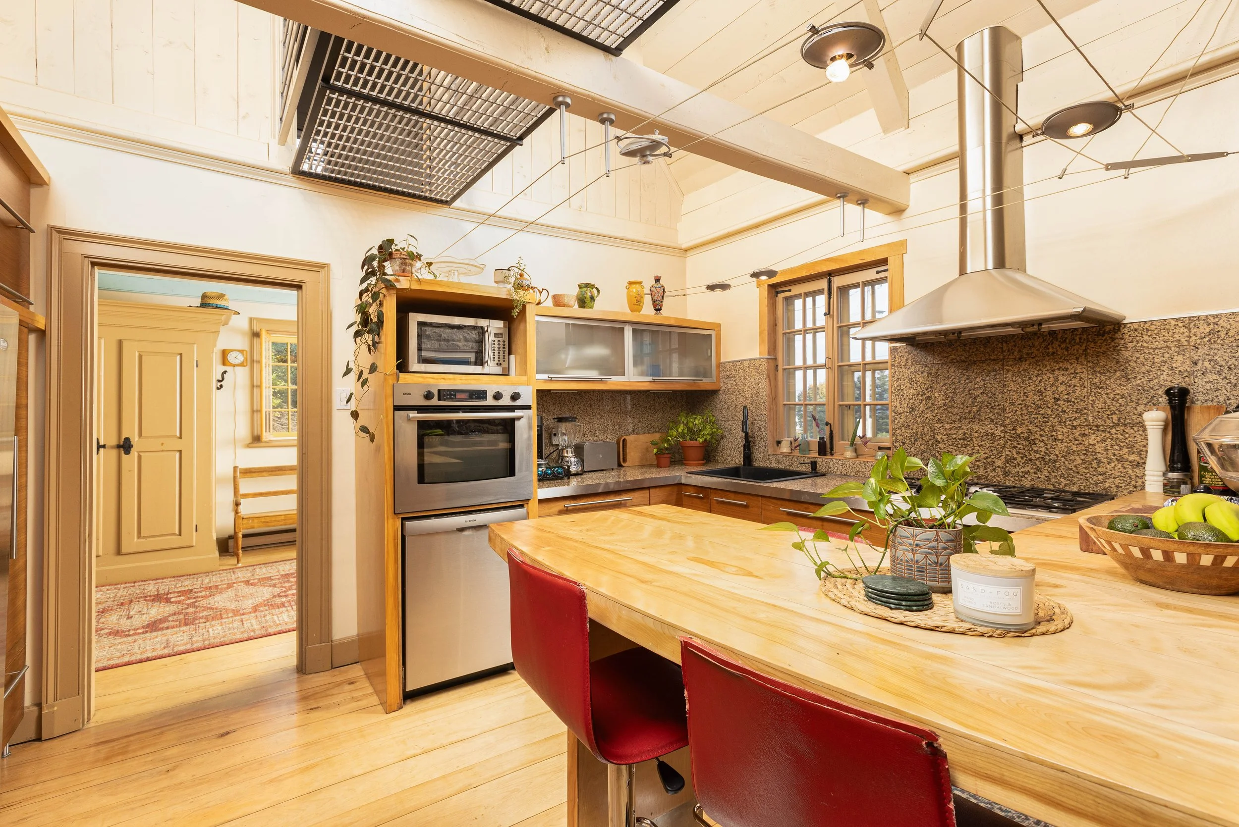 Cuisine rustique avec plan de travail en bois, fenêtres à petits carreaux, étagères en bois, plantes vertes et appareils en acier inoxydable.