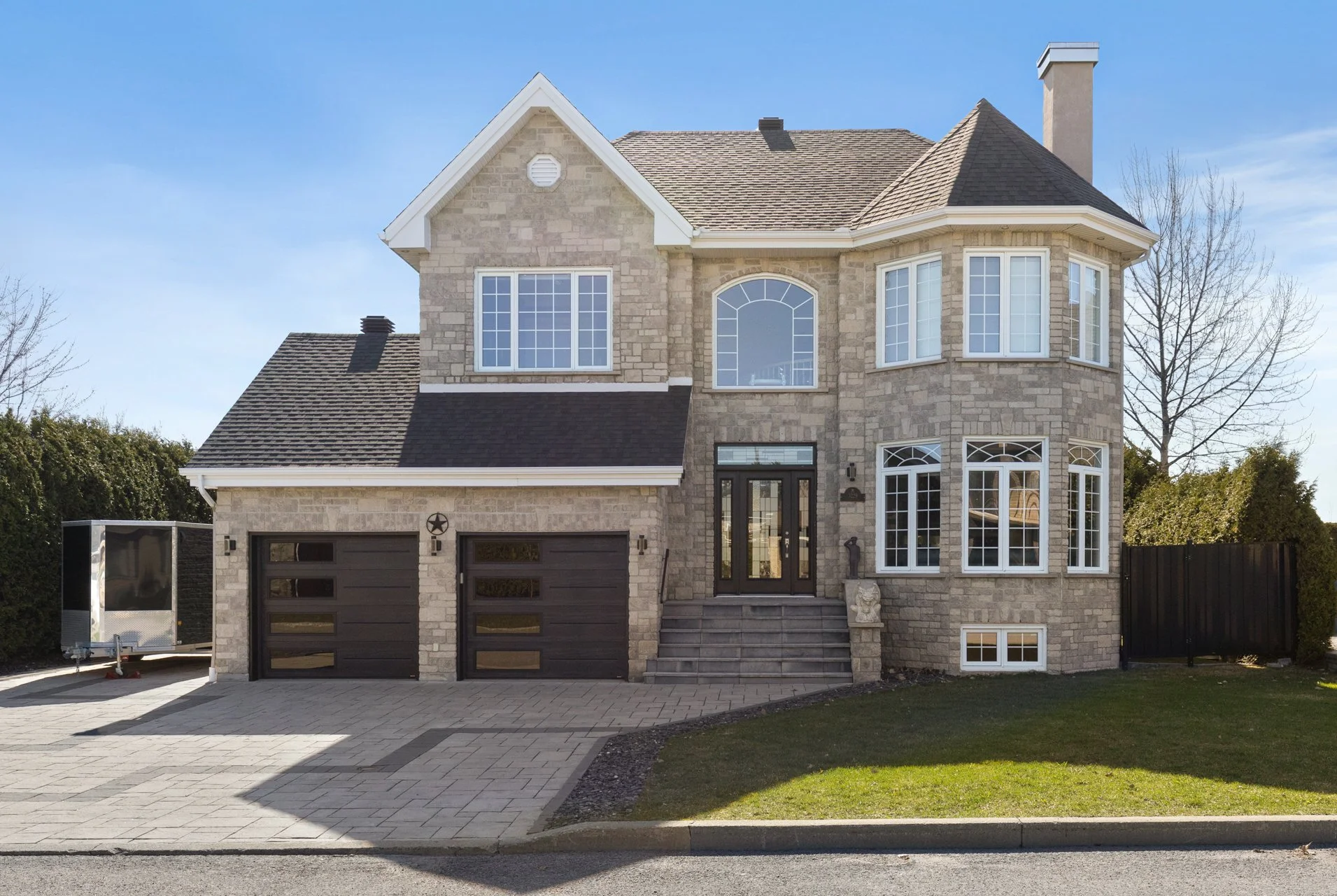 Maison moderne à plusieurs étages avec façade en pierre, fenêtres grand format et porte d'entrée en verre, devant un jardin et un chemin pavé.