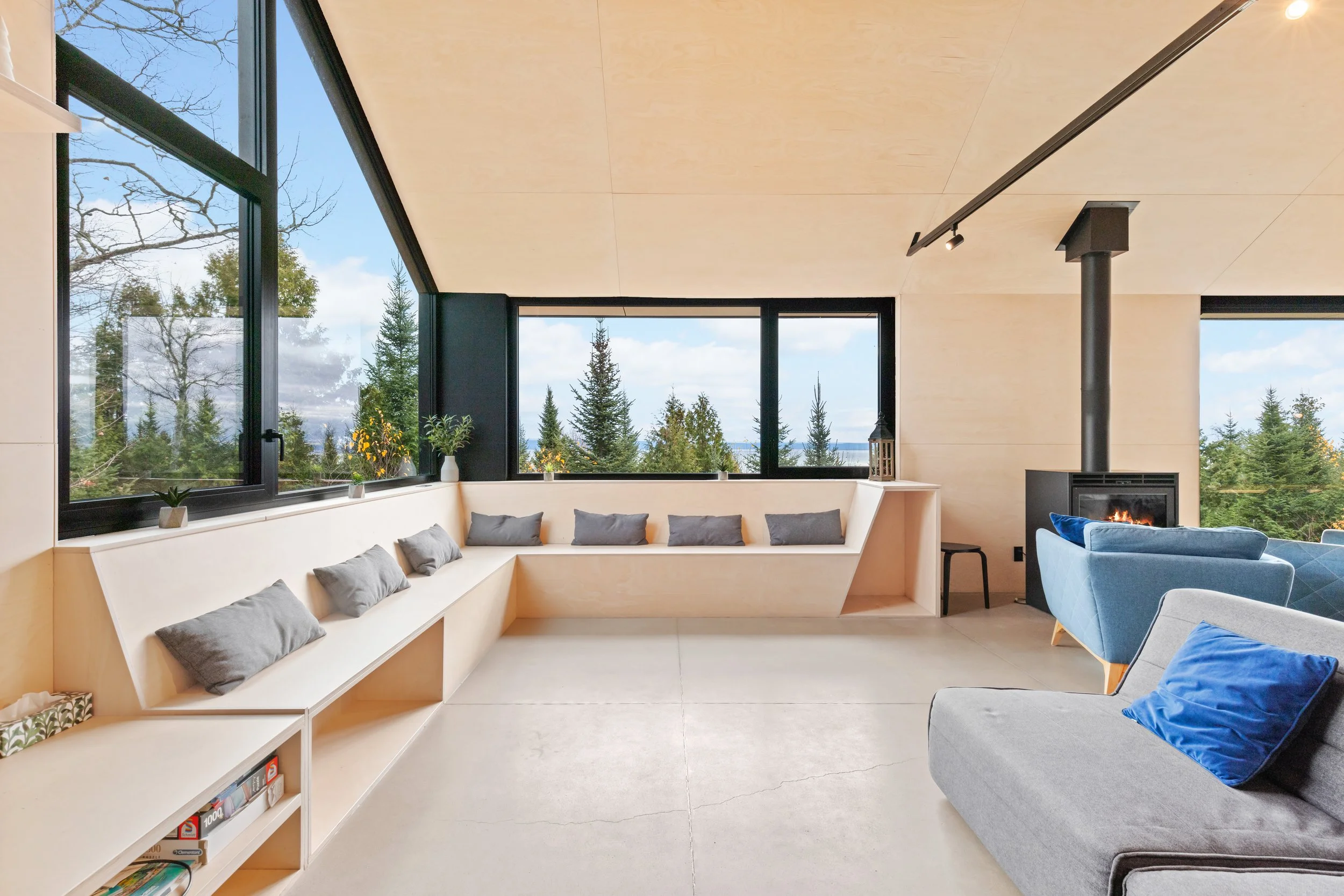 Salon moderne avec de grandes fenêtres offrant une vue sur la forêt, canapés bleus et gris, cheminée au bois, décor minimaliste.