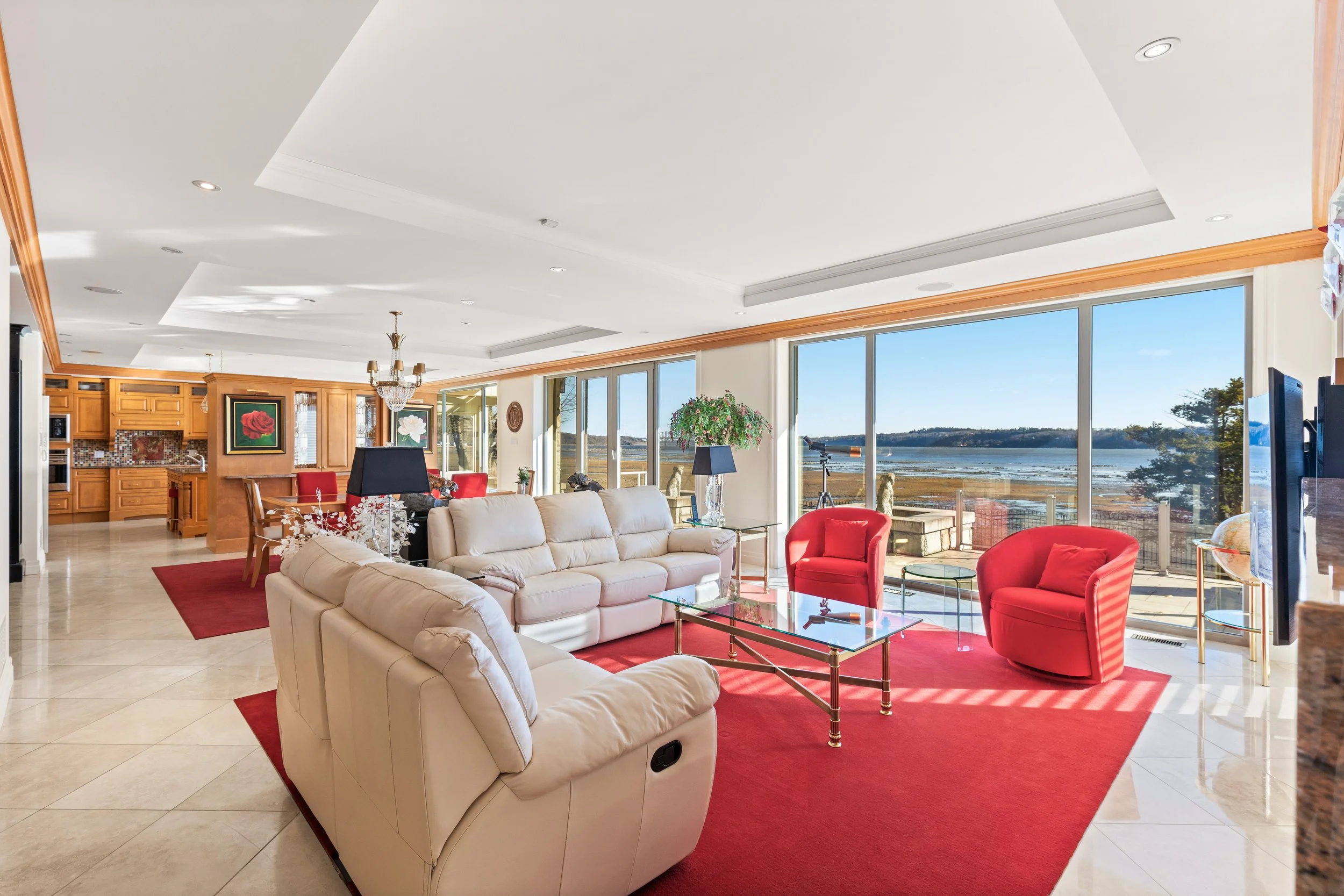 Salon spacieux avec grandes fenêtres offrant une vue sur l'extérieur, comprenant des canapés blancs, deux fauteuils rouges, tapis rouge, table en verre avec cadre doré, décorations florales, et une vue sur un paysage marin.