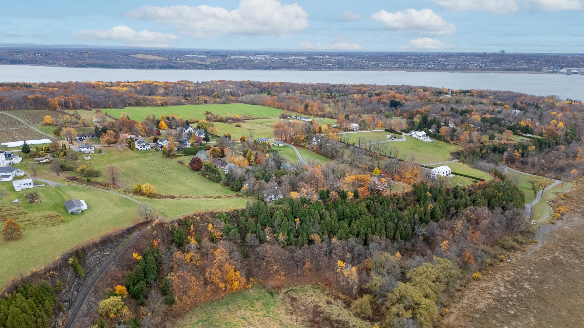 Vallée offrant une vue aérienne avec des maisons réparties sur des terrains verdoyants, entourée de bois, avec une rivière en arrière-plan et un ciel nuageux.