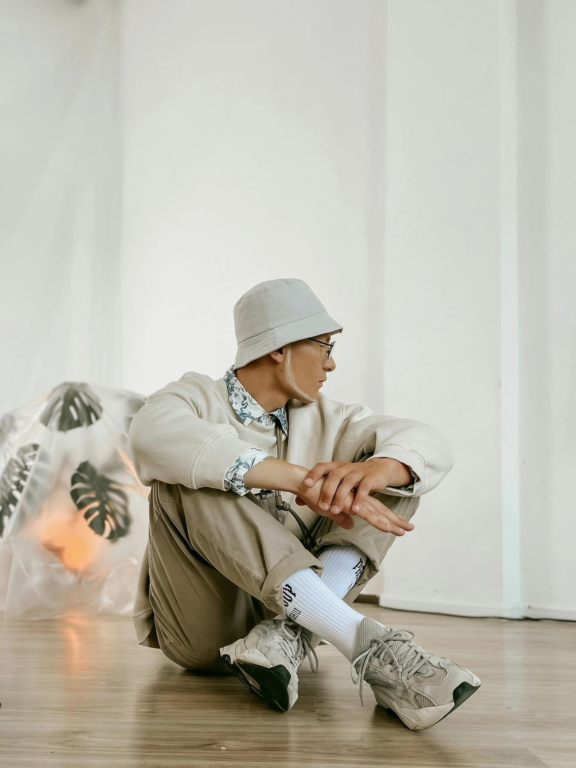 Person wearing beige bucket hat, glasses, beige sweatshirt, khaki pants, white socks, and sneakers, sitting on the wooden floor with arms resting on knees, in a minimalist indoor space.