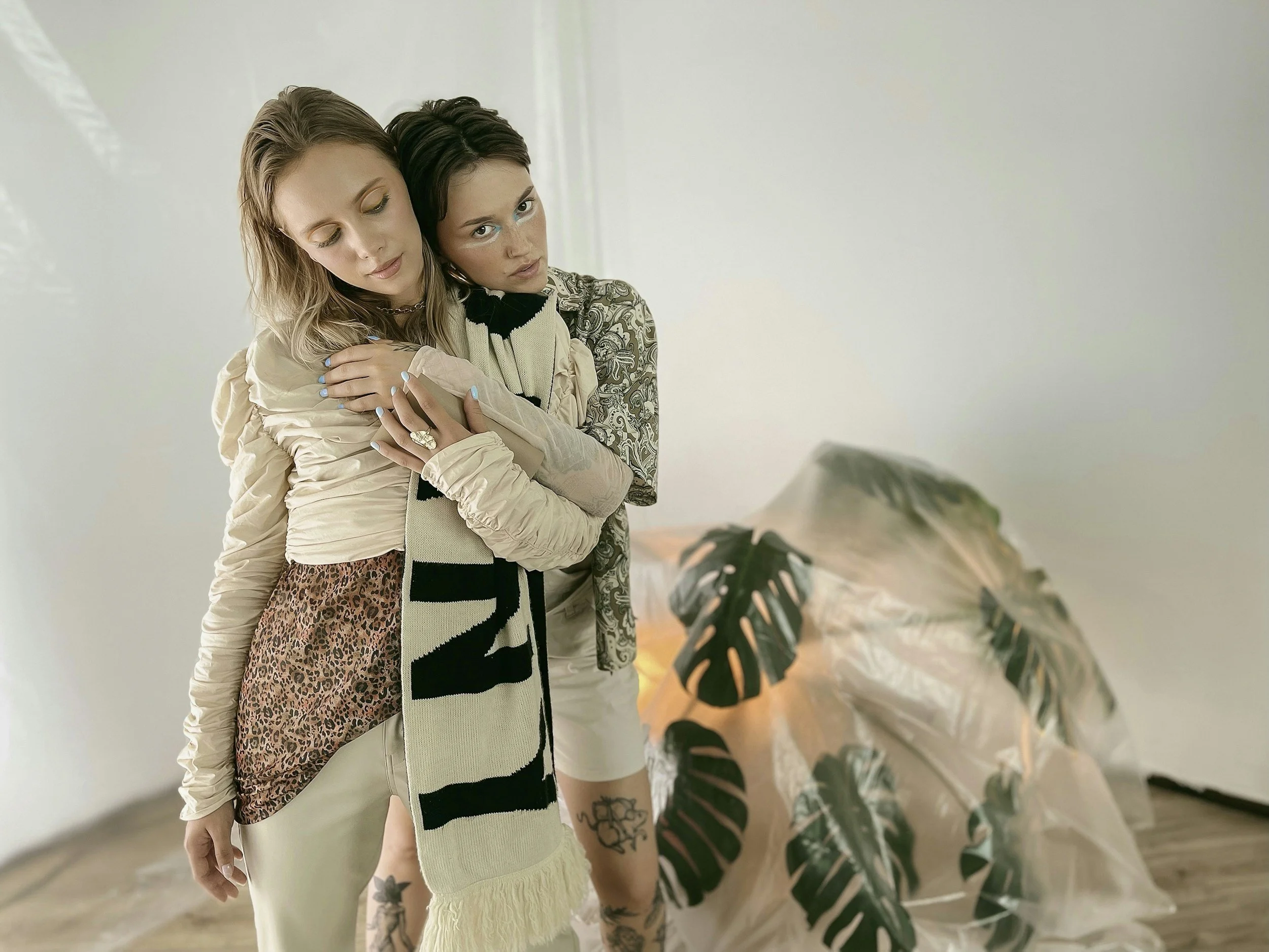 Two women embracing in front of a white wall and a large leafy plant.