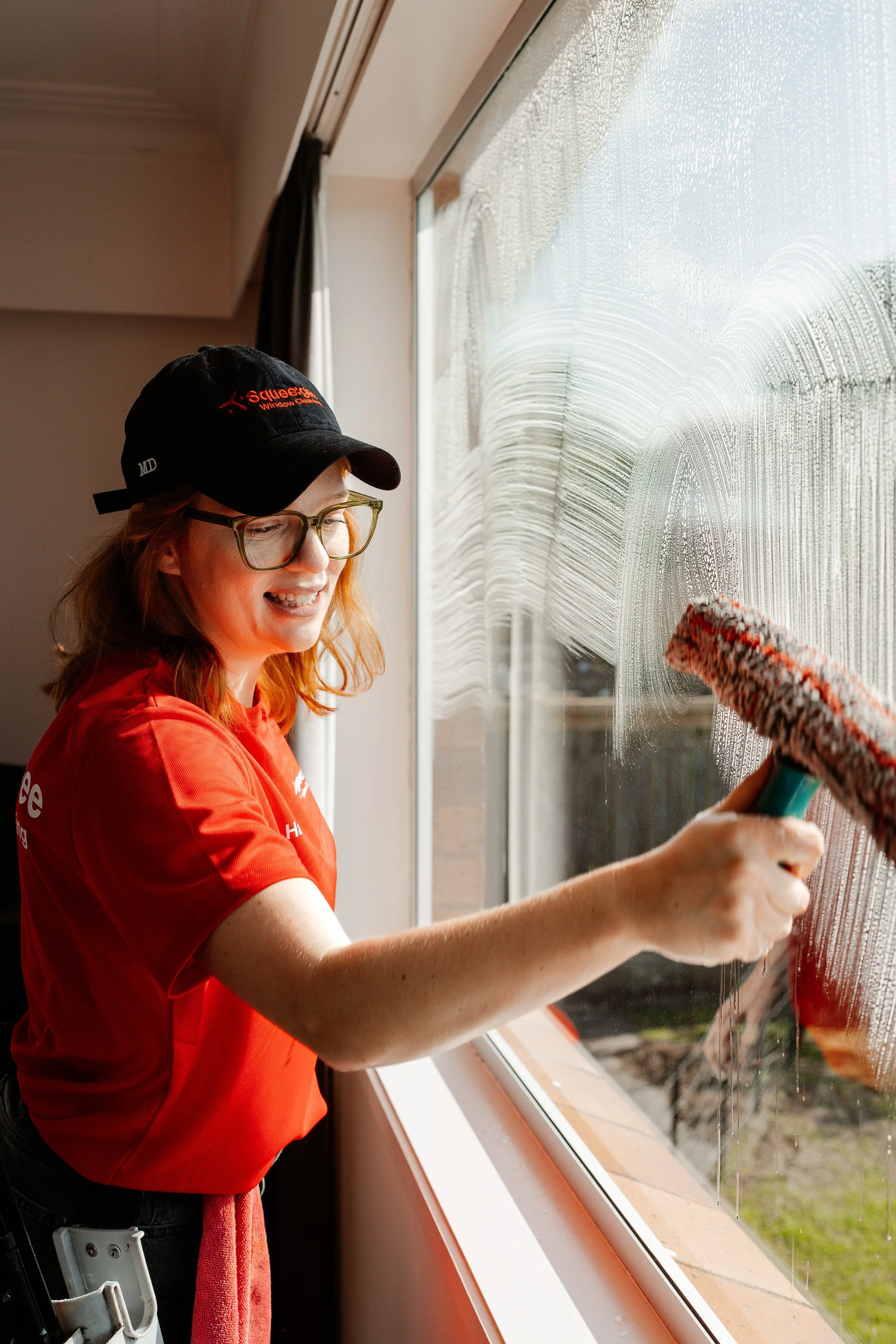 A woman cleaning a large window with a cloth and spray bottle, wearing a black cap and red shirt, with cleaning tools attached to her waist.