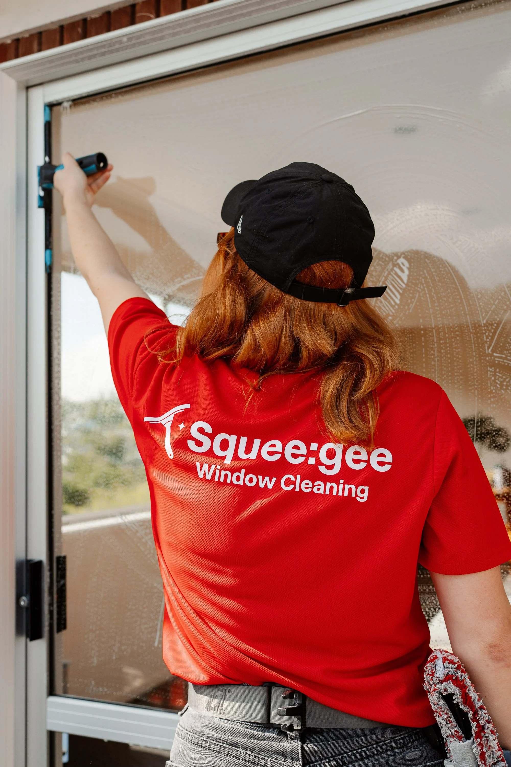 A person cleaning a window with a squeegee. They are wearing a red shirt with white text that reads 'T Squee:gee Window Cleaning,' a black cap, gray pants, and a patterned cloth hanging from their pocket.