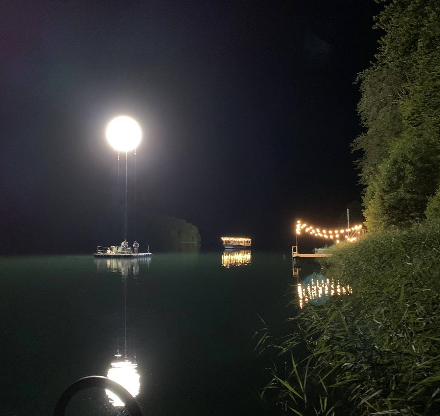 Nachtbild eines Sees mit einer Ponton, einem Balloon Light darüber, einer beleuchteten Brücke und Bäume am Ufer