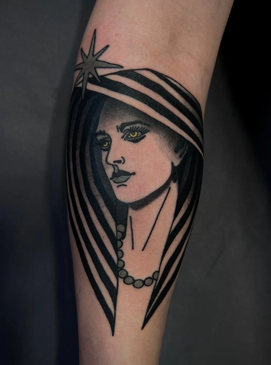Tattoo of a woman with yellow eyes, dark hair, and a beaded necklace, wearing a hat with a star.