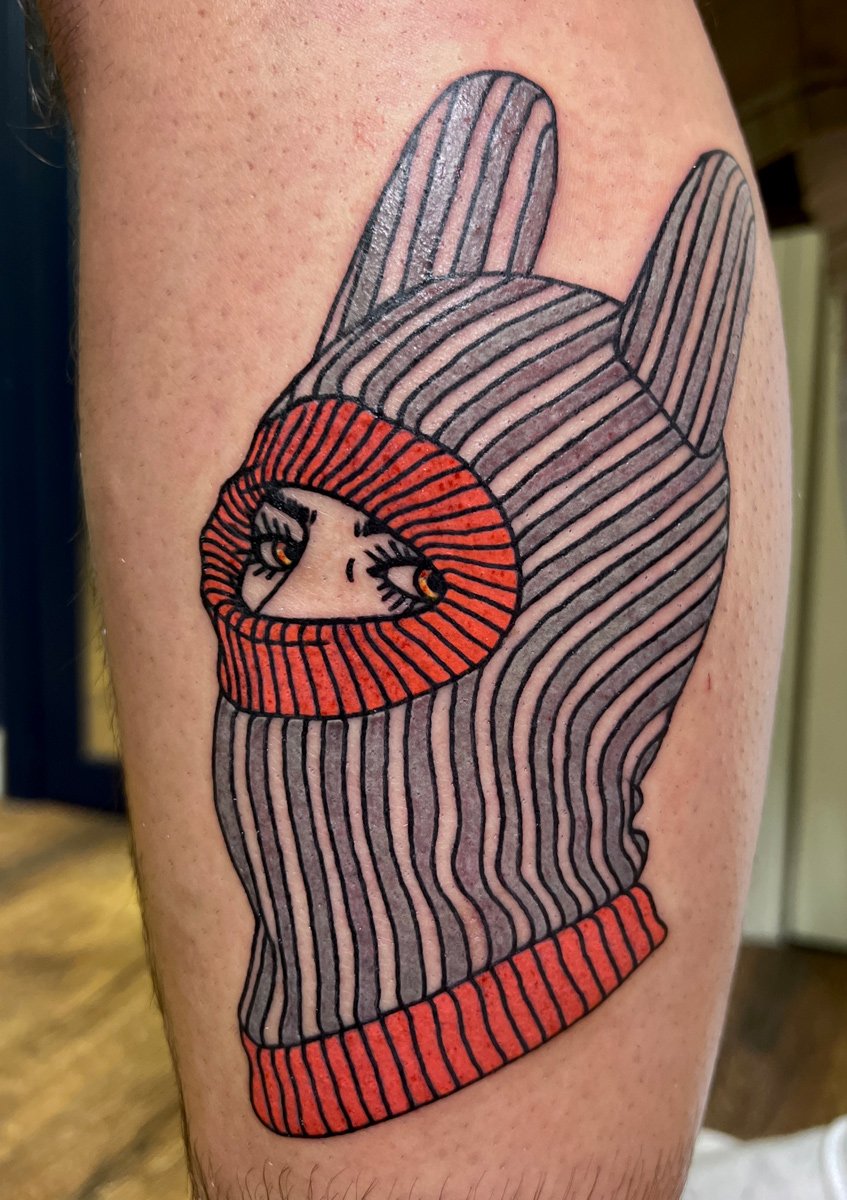 Tattoo of a mitten character with a face and eyes, wearing a striped hat and scarf, on a person's arm.