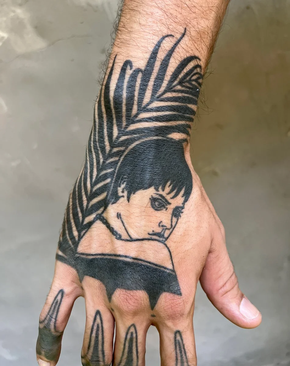 Tattoo of a girl's face with short hair and a feathered headdress on a person's hand.