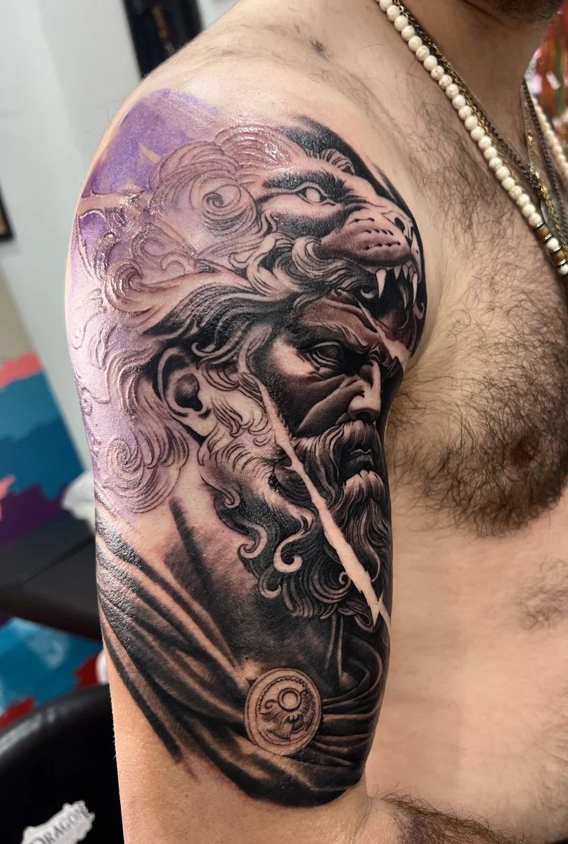 A black and gray tattoo on a man's upper arm depicts a detailed image of a bearded man with long hair, holding a pipe in his mouth, with a lion's head above him and a circular emblem below.