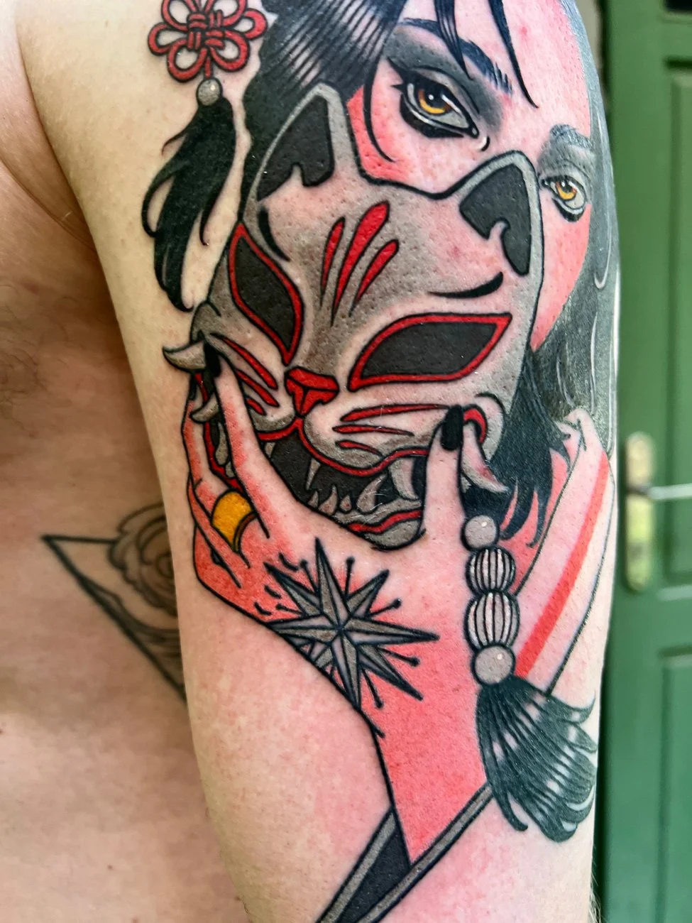 Tattoo of a woman with black hair and yellow eyes, along with a stylized cat face with black, red, and pink accents, and a star-shaped tattoo below. The woman is holding her finger to her lips in a shushing gesture.