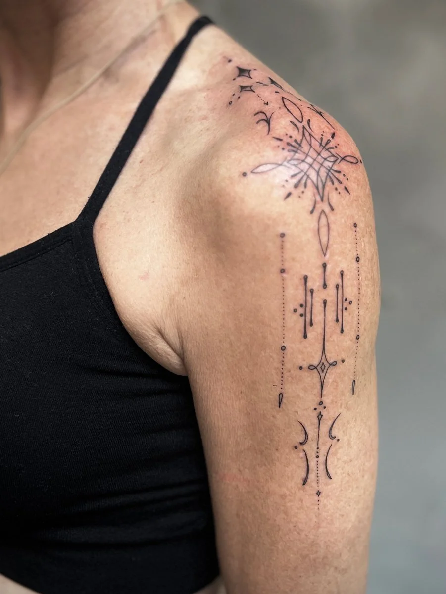 Close-up of a person's upper arm with a minimalist black tattoo featuring geometric and celestial elements on the shoulder and upper arm.