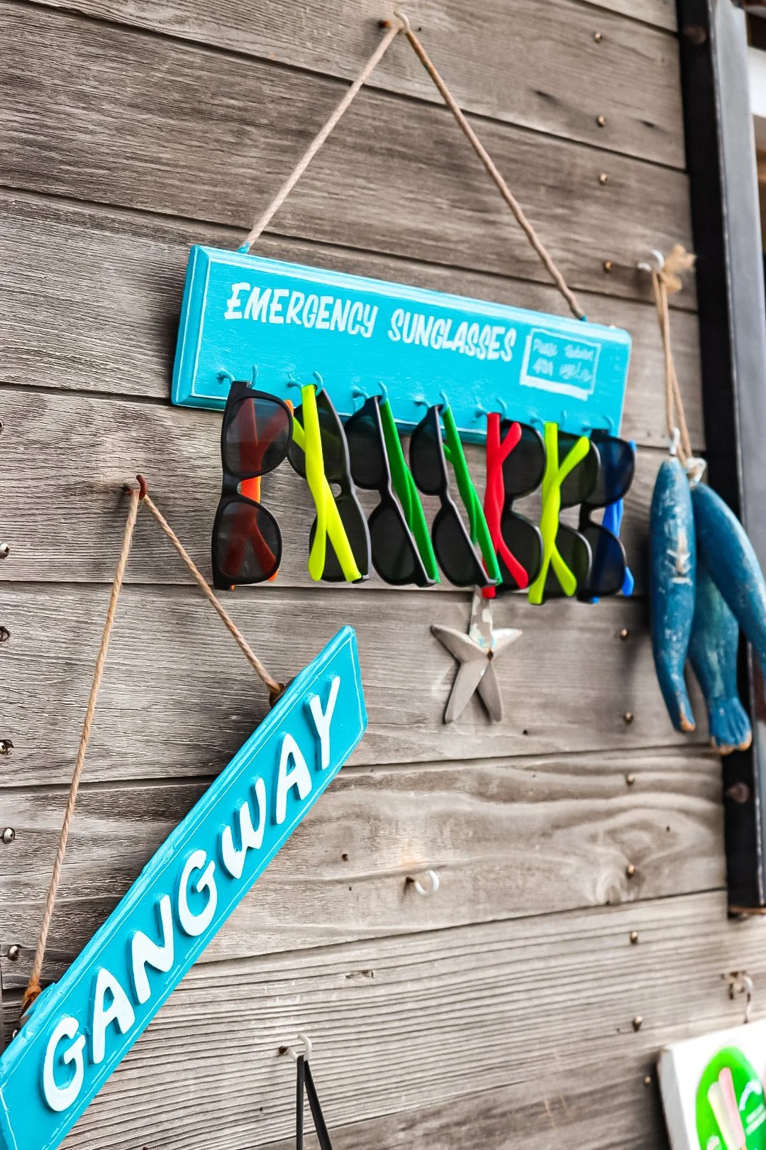 A wooden wall displays a blue sign labeled 'Emergency Sunglasses' with a line of sunglasses hanging from hooks in front of it, and a blue sign with 'GANGUAY' on it hanging by a rope.