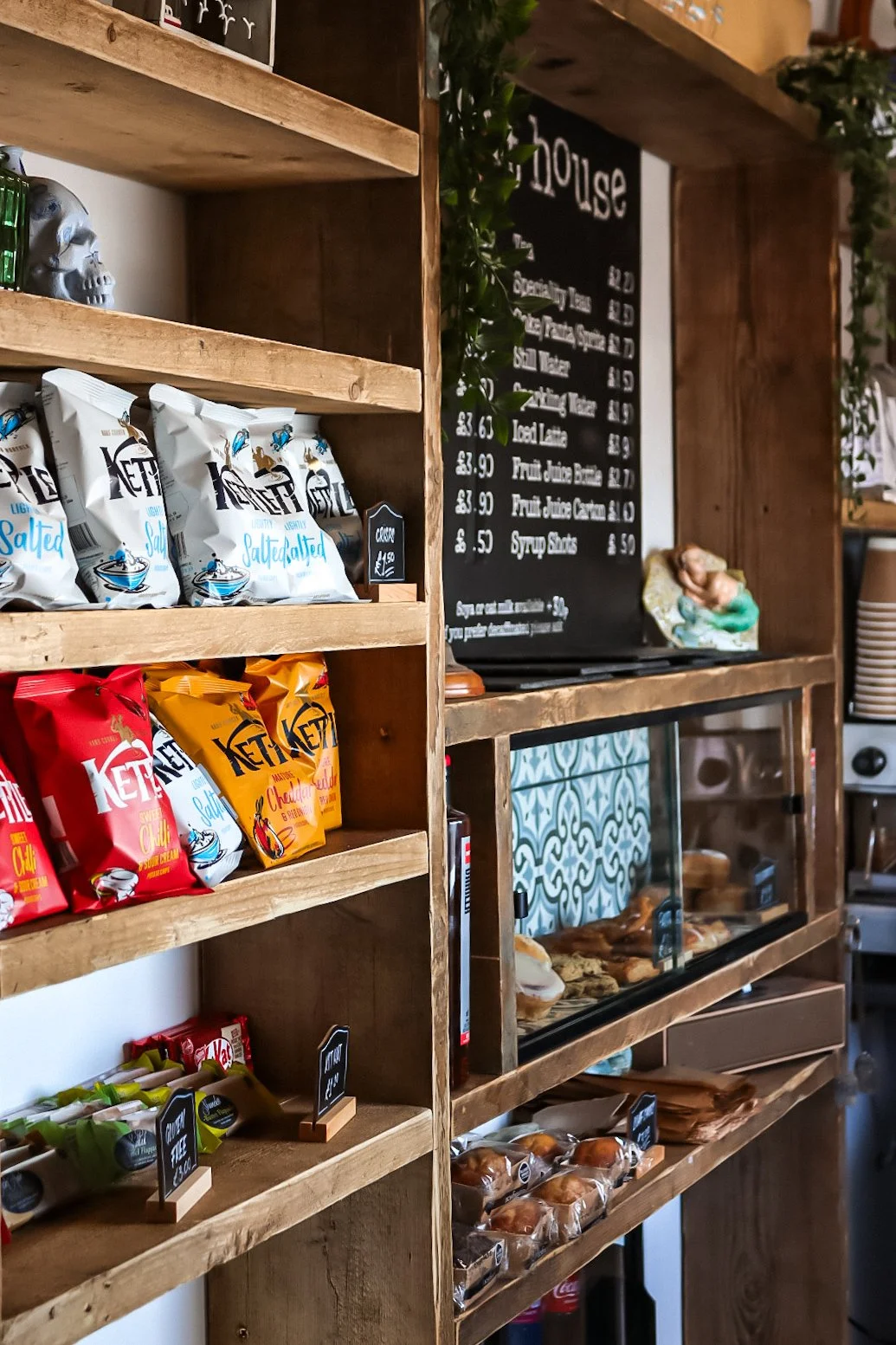 Wooden shelves with packets of salt and chips in a cafe or bakery. A blackboard menu and baked goods behind a glass display case are visible in the background.