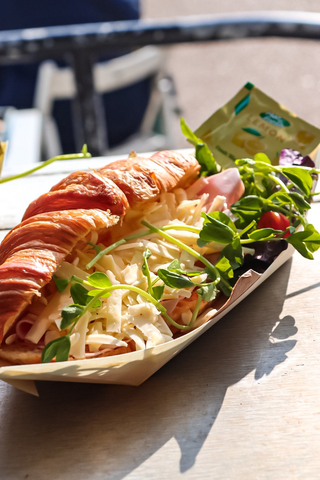 A tray of assorted cold cuts, shredded cheese, fresh greens, cherry tomatoes, and a croissant on a wooden table outdoors.