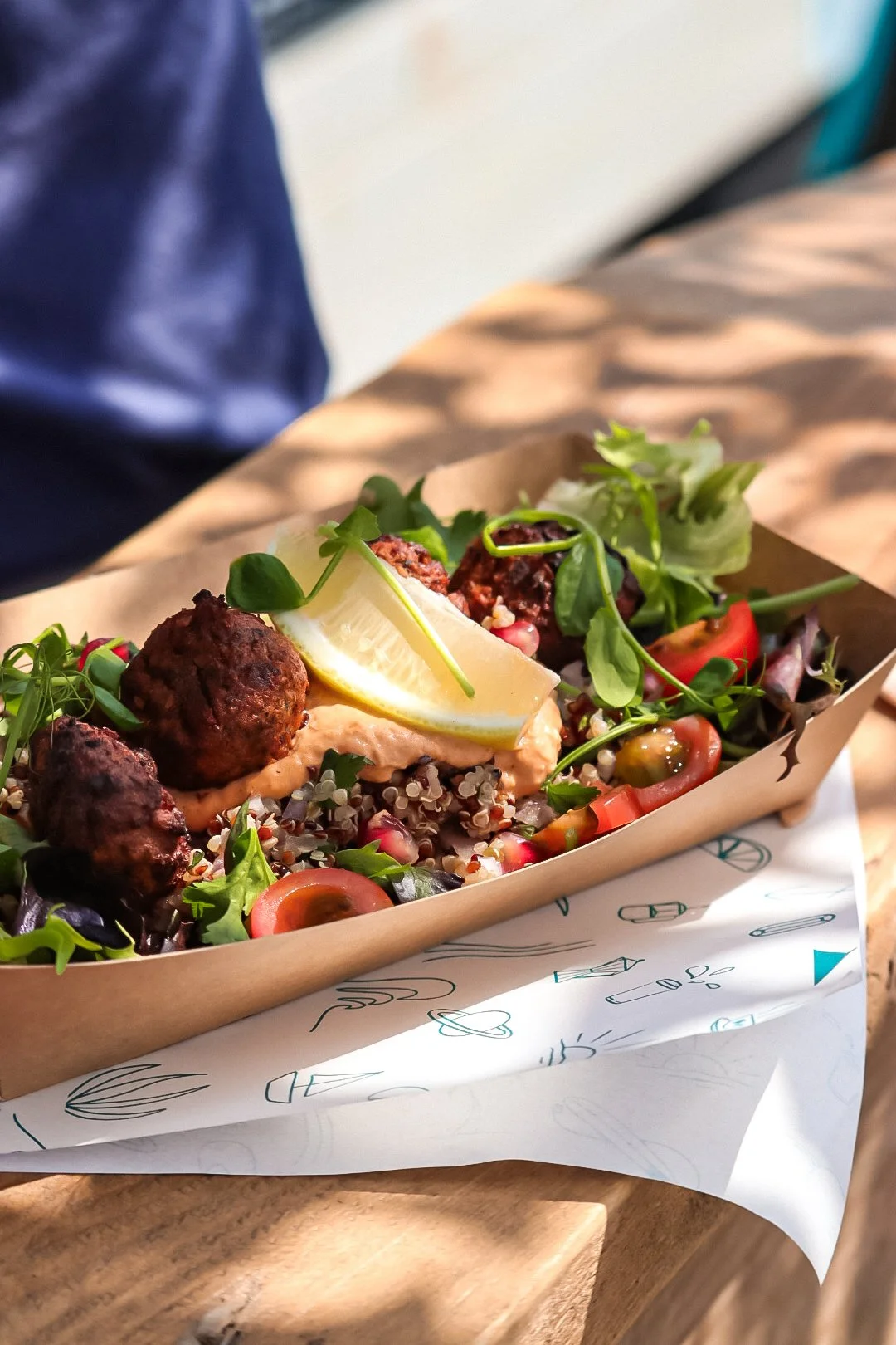 A serving tray of mixed salad with falafel balls, cherry tomatoes, lemon wedge, and greens on paper-lined wooden tray.