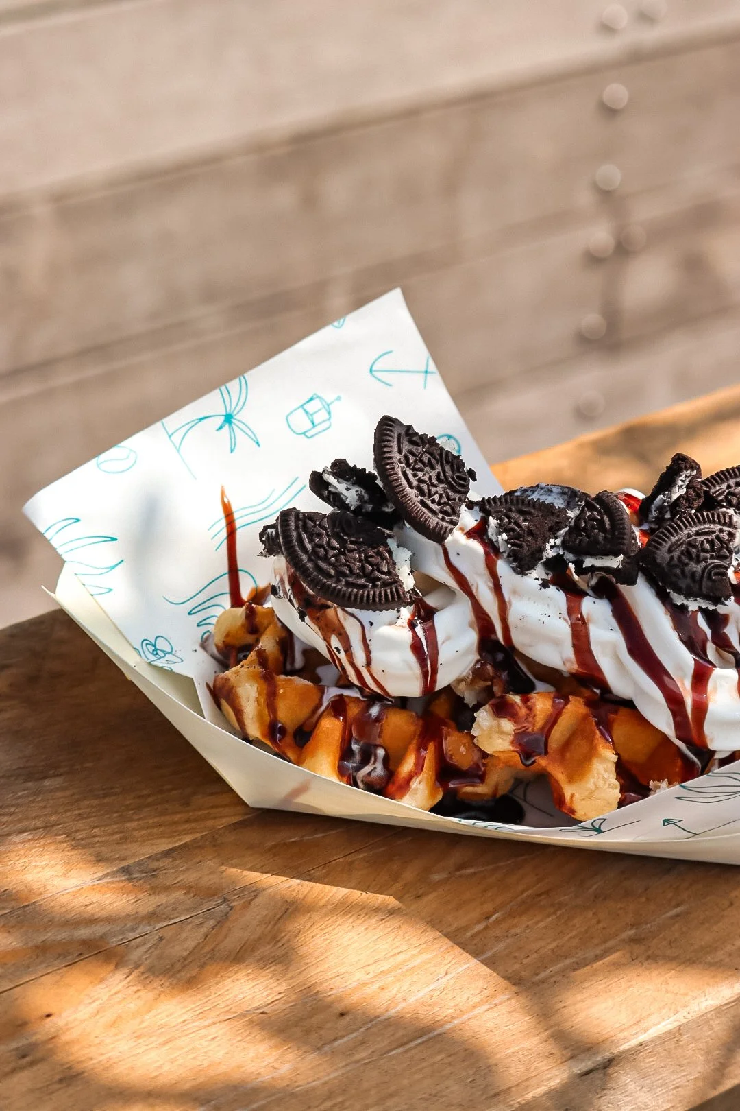 Waffle topped with whipped cream, Oreo cookies, chocolate syrup, and caramel sauce in a paper tray on a wooden table.