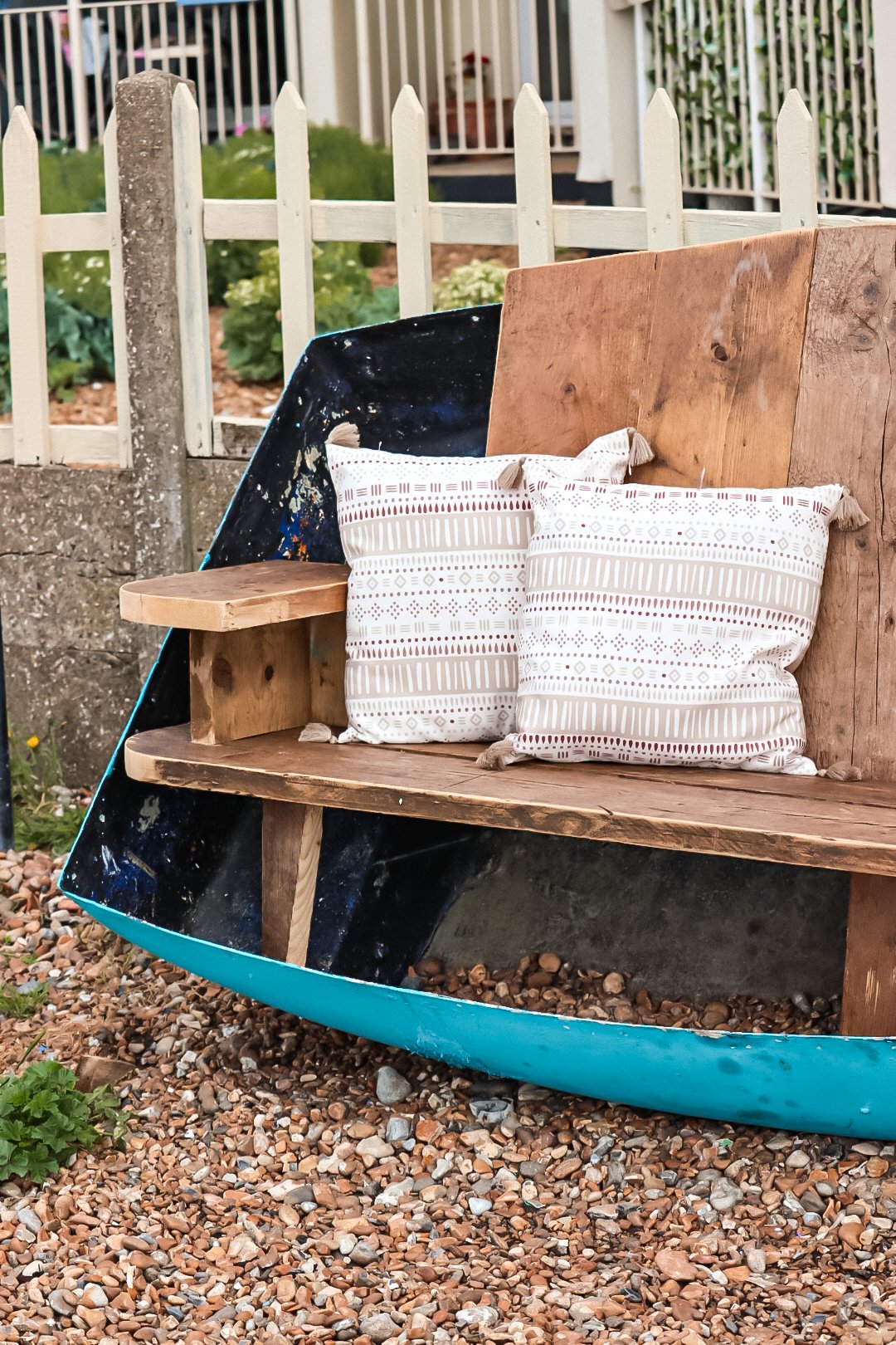 A boat repurposed as a bench with two decorative pillows, set on a gravel surface, in front of a white fence and garden.