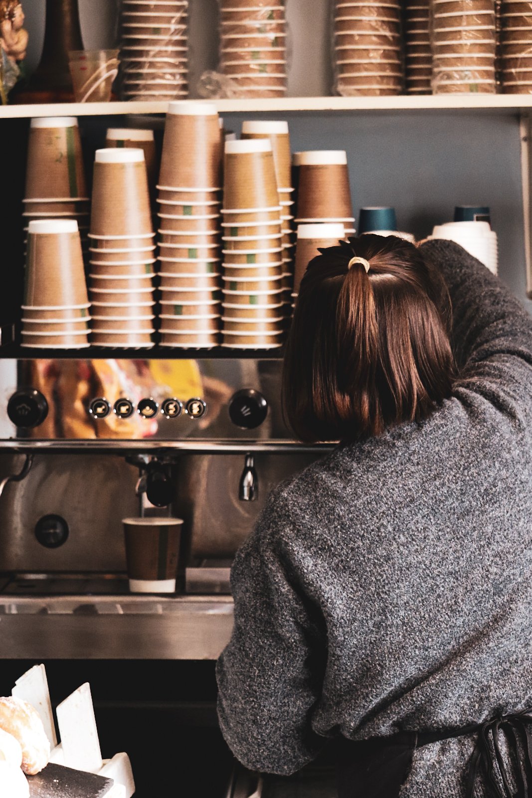 A person with short brown hair and a gray coat preparing coffee at a coffee shop, with stacked paper cups on a shelf behind them and a coffee machine in front.
