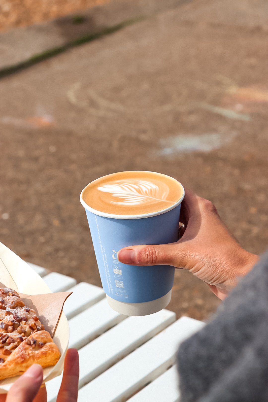 A person holding a blue paper cup of latte with latte art featuring a leaf pattern, next to a plate of pastries, outdoors on a white bench.